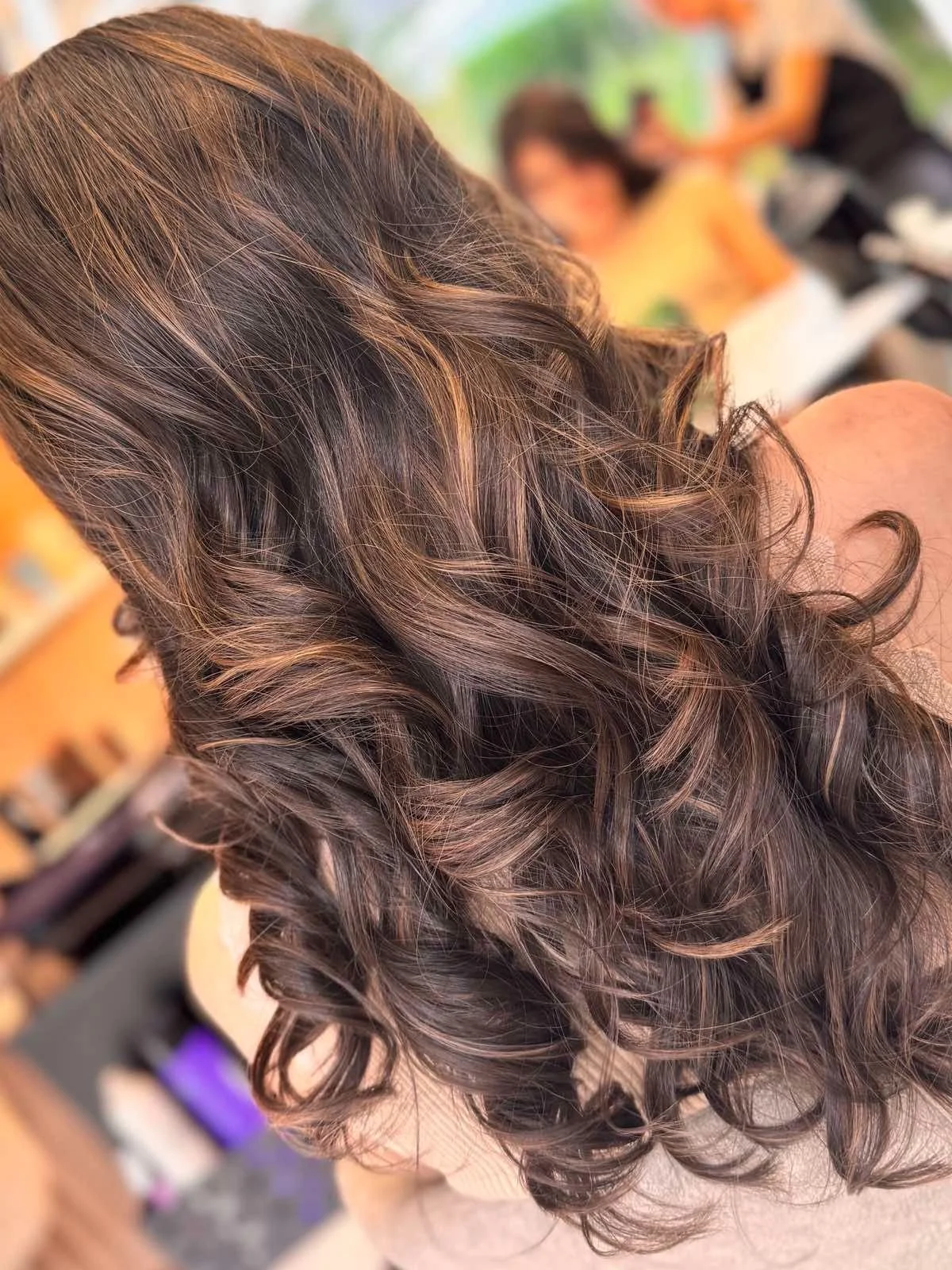 Close-up of a woman's long, wavy, brunette hair with highlights, with out-of-focus people in the background.