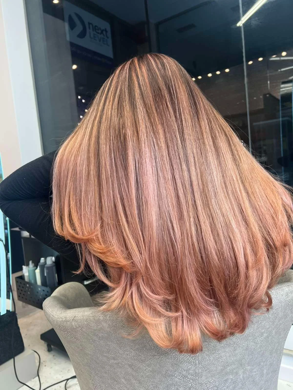 Back view of a woman with long, smooth, rose gold and blonde ombre hair sitting in a salon chair.