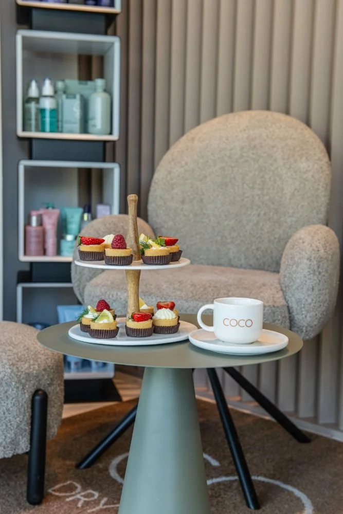 A small round table with a two-tier serving tray of assorted cupcakes and a cup of coffee labeled 'COCO' in a cozy room.