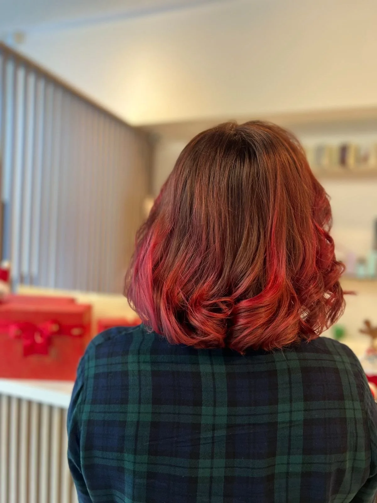 Back of woman with shoulder-length brown hair with red highlights and curling, wearing a dark plaid shirt, in a room with holiday decorations.