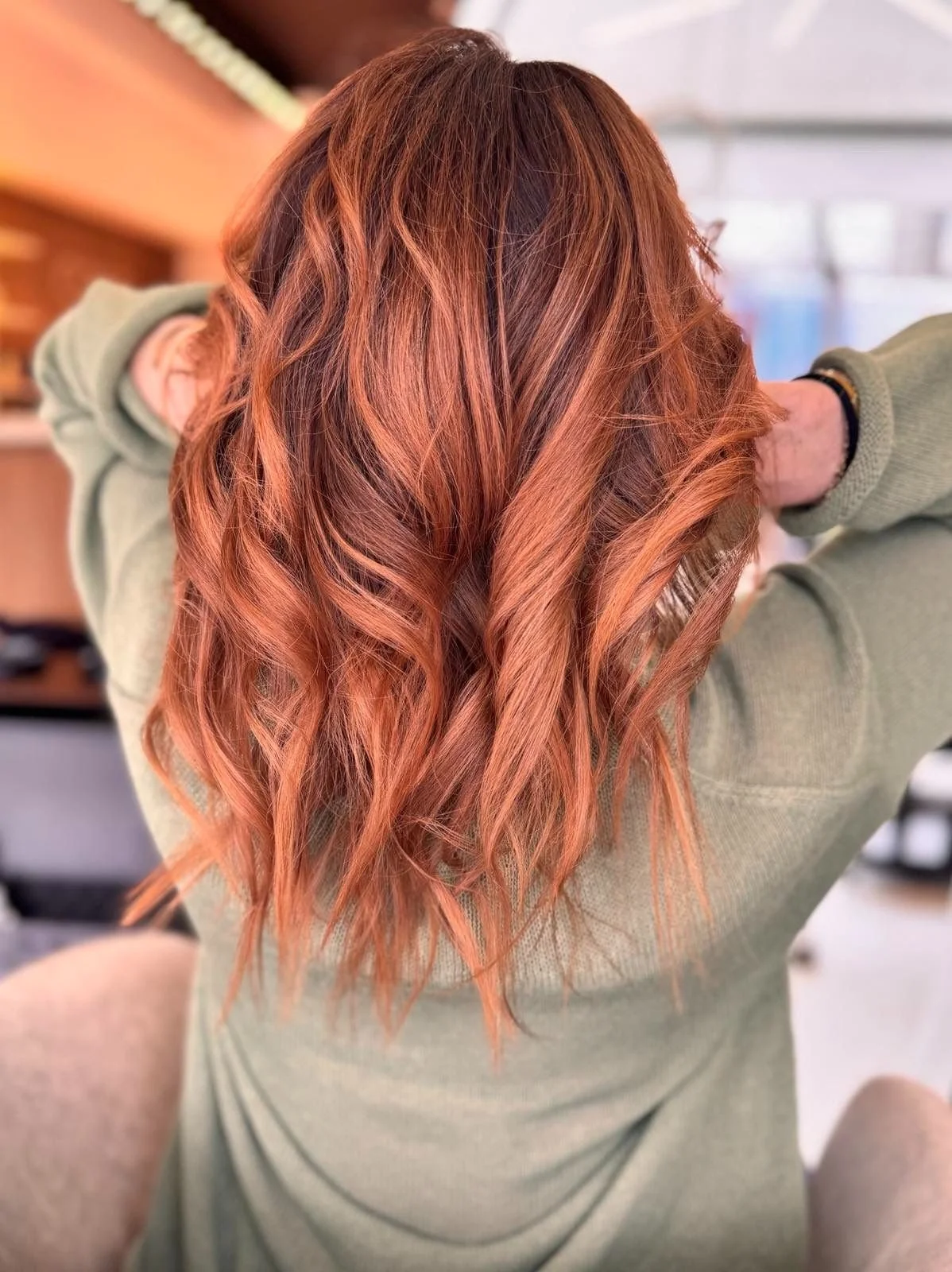 Back view of a woman with long, wavy, reddish-orange hair, wearing a green hoodie, with her arms raised behind her head.