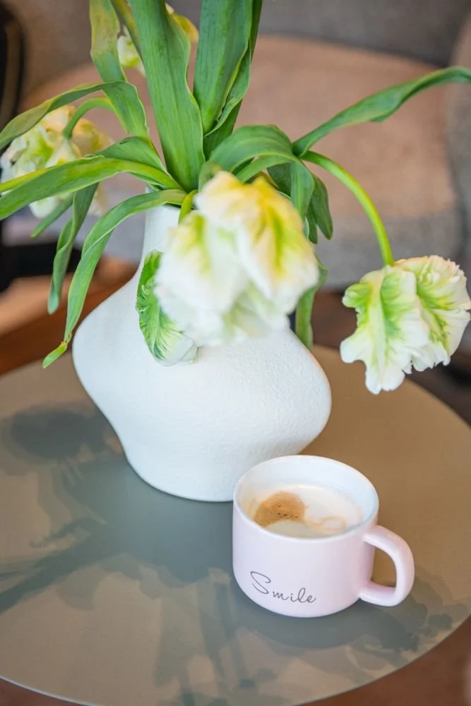 A white vase with green leaves and white and green flowers on a gray table. A small pink mug with coffee and the word 'Smile' written on it is in front of the vase.