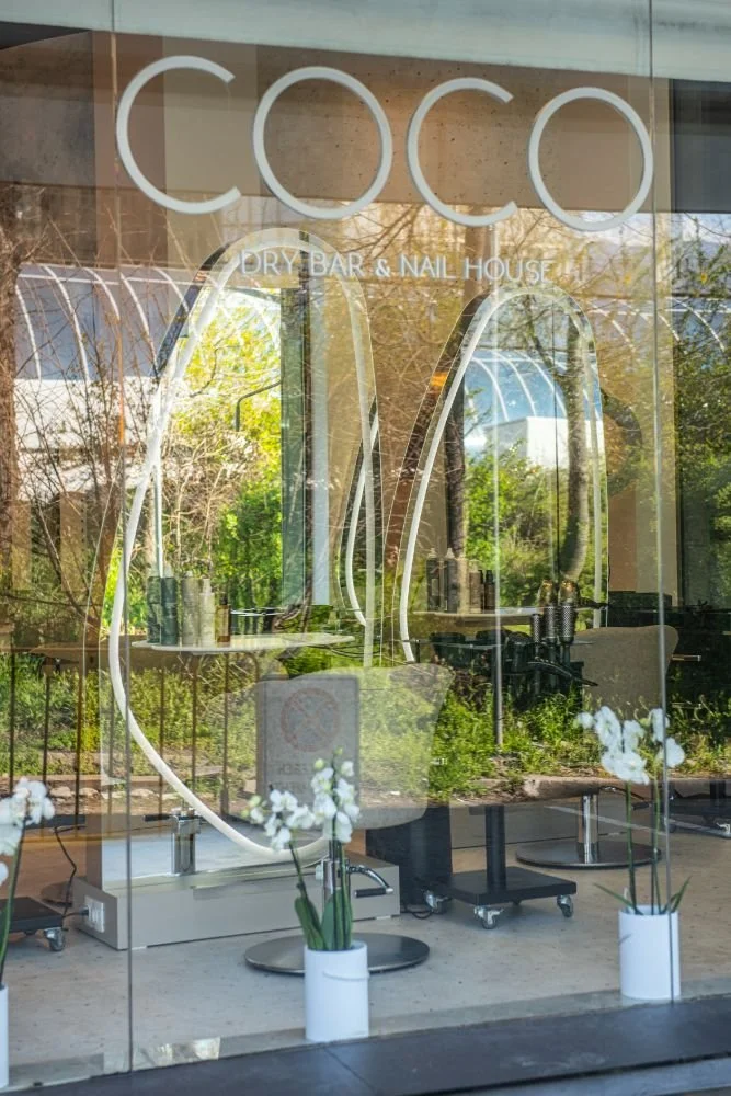 The storefront window displays a sign that reads 'COCO' and 'DRY BAR & NAIL HOUSE' underneath. Inside, there are styling chairs, mirrors, and hair or nail salon equipment, with reflections of trees and outdoor scenery visible through the glass.