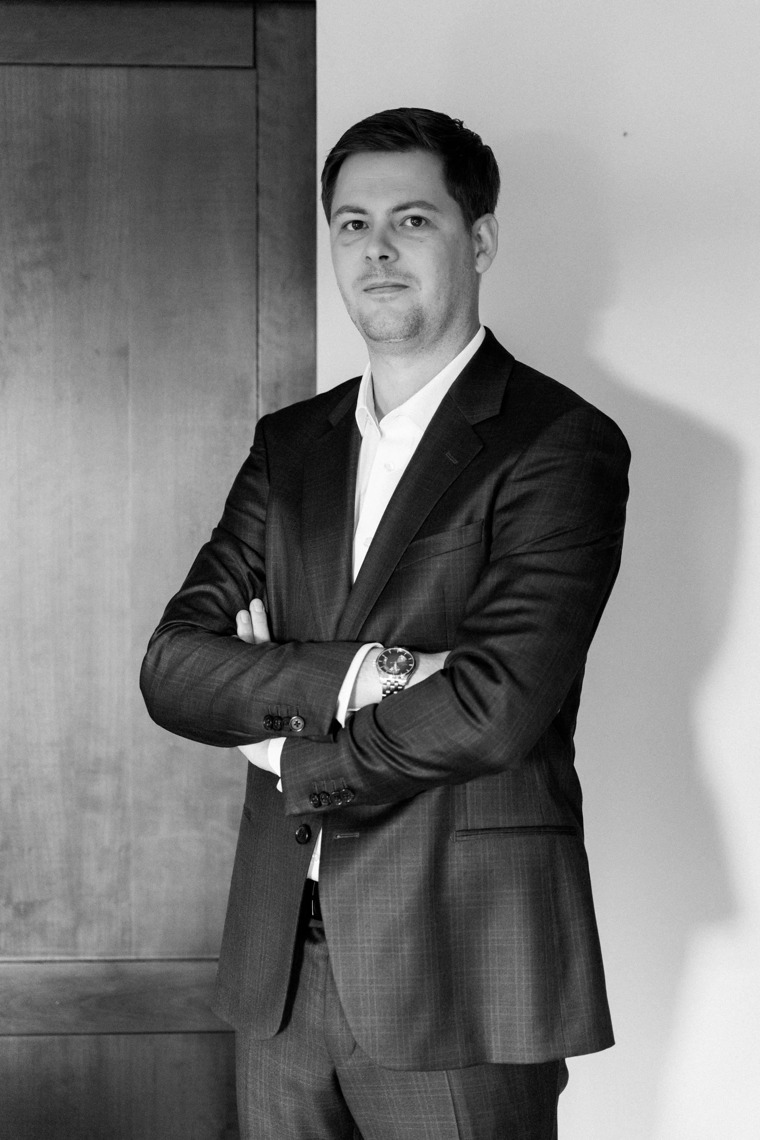 Zwart-wit foto van een man in een pak met gezichtsuitdrukking en armen over elkaar, staand binnen, met een houten wand en witte muur als achtergrond.