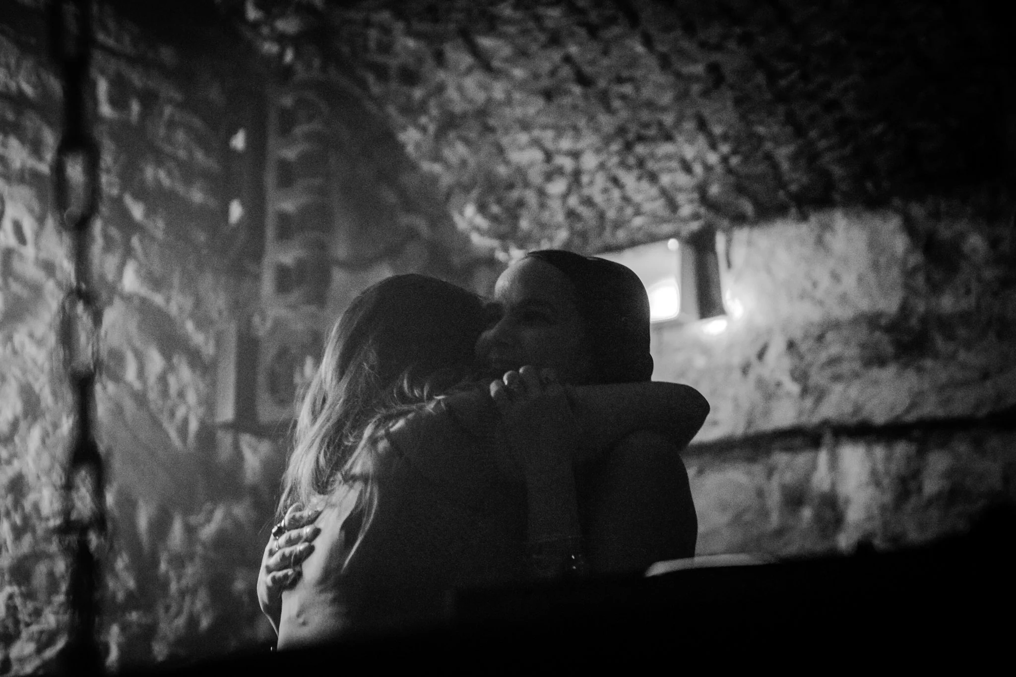 Silhouette of a couple hugging and kissing in a dimly lit, rustic interior with stone walls and small window in the background.