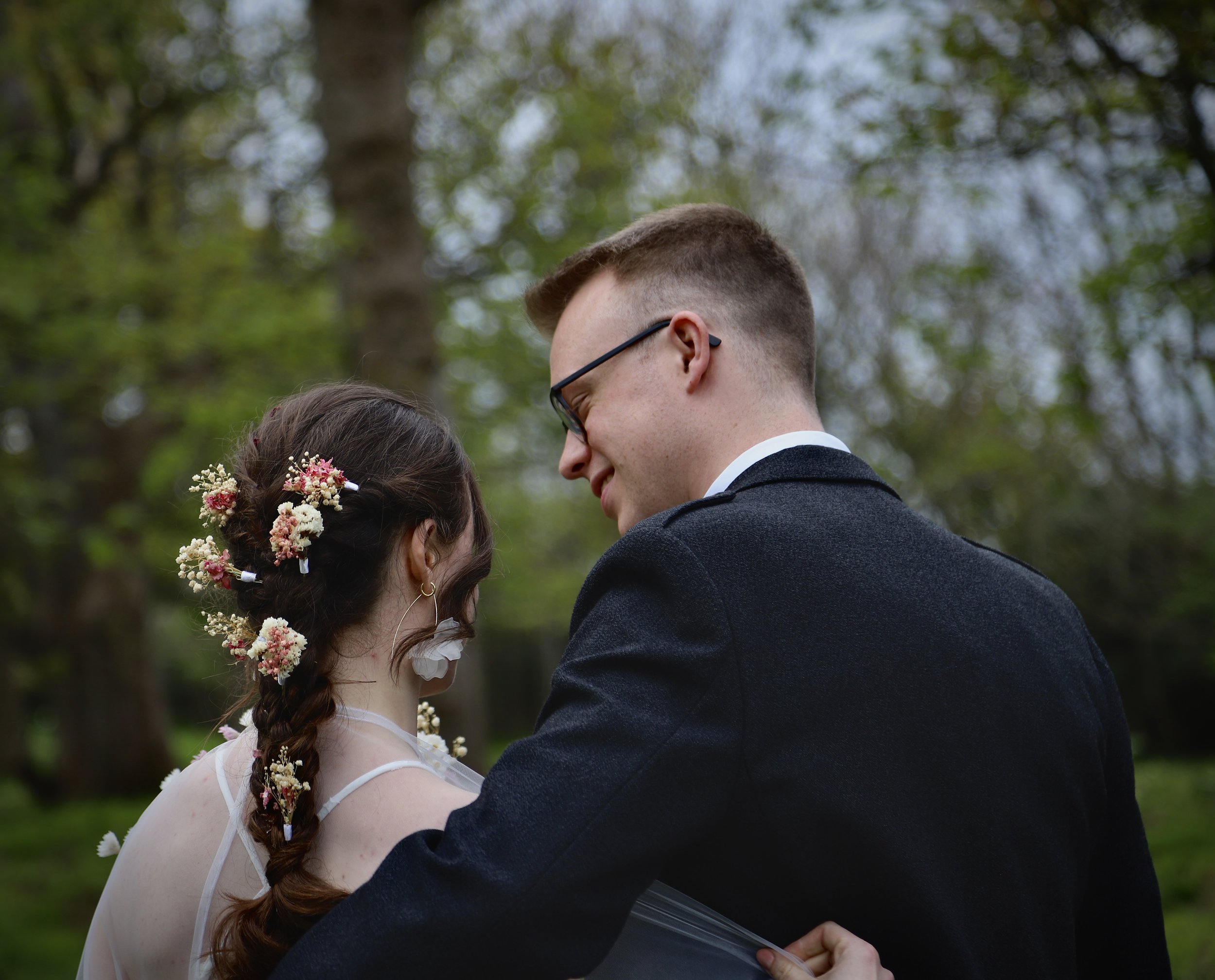A wedding couple is hugging outdoors with trees in the background, the woman has flowers in her hair and is wearing a white dress, and the man is wearing a dark suit and glasses.
