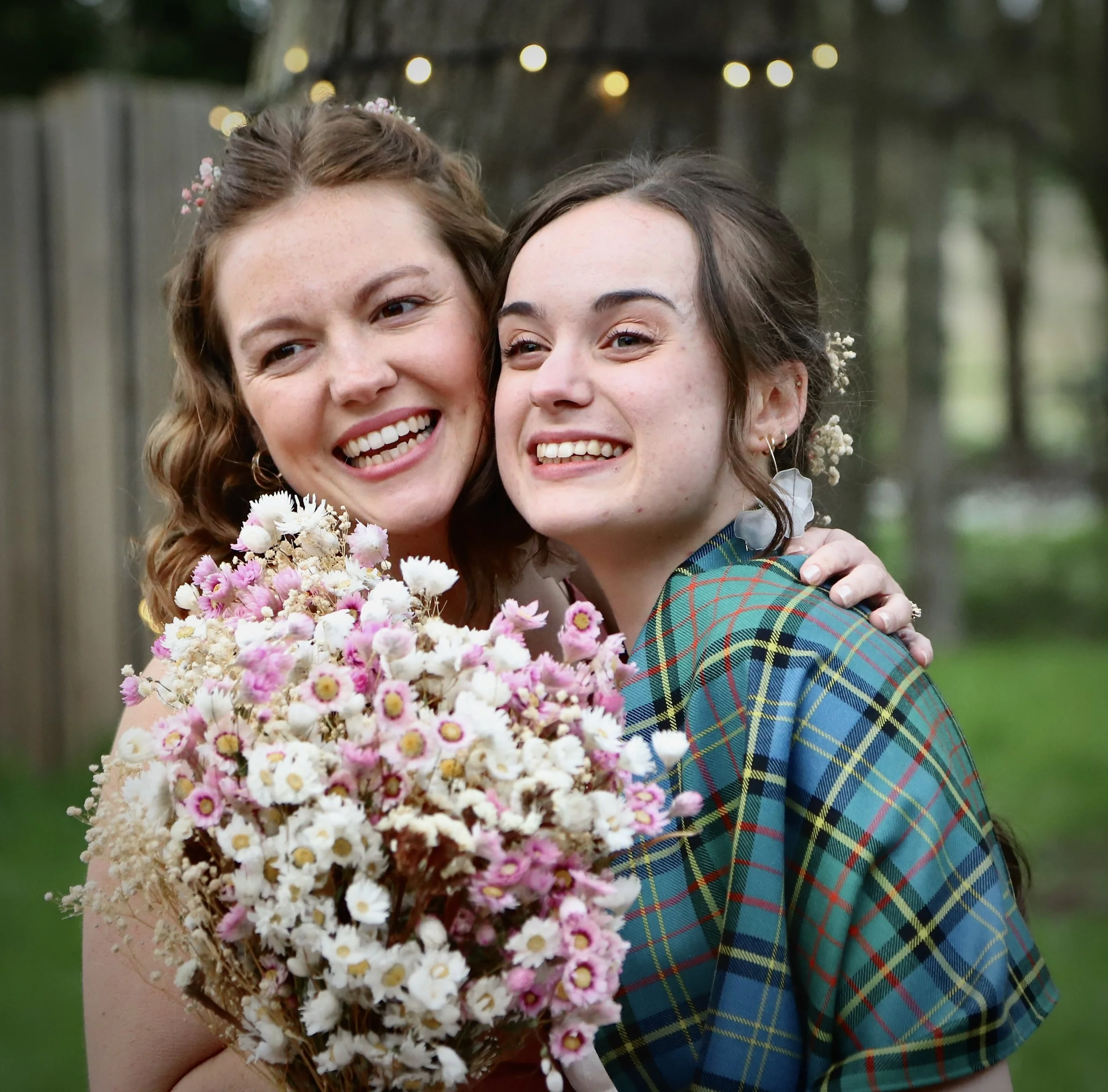 Two women smiling and hugging outdoors, one holding a bouquet of pink and white flowers.