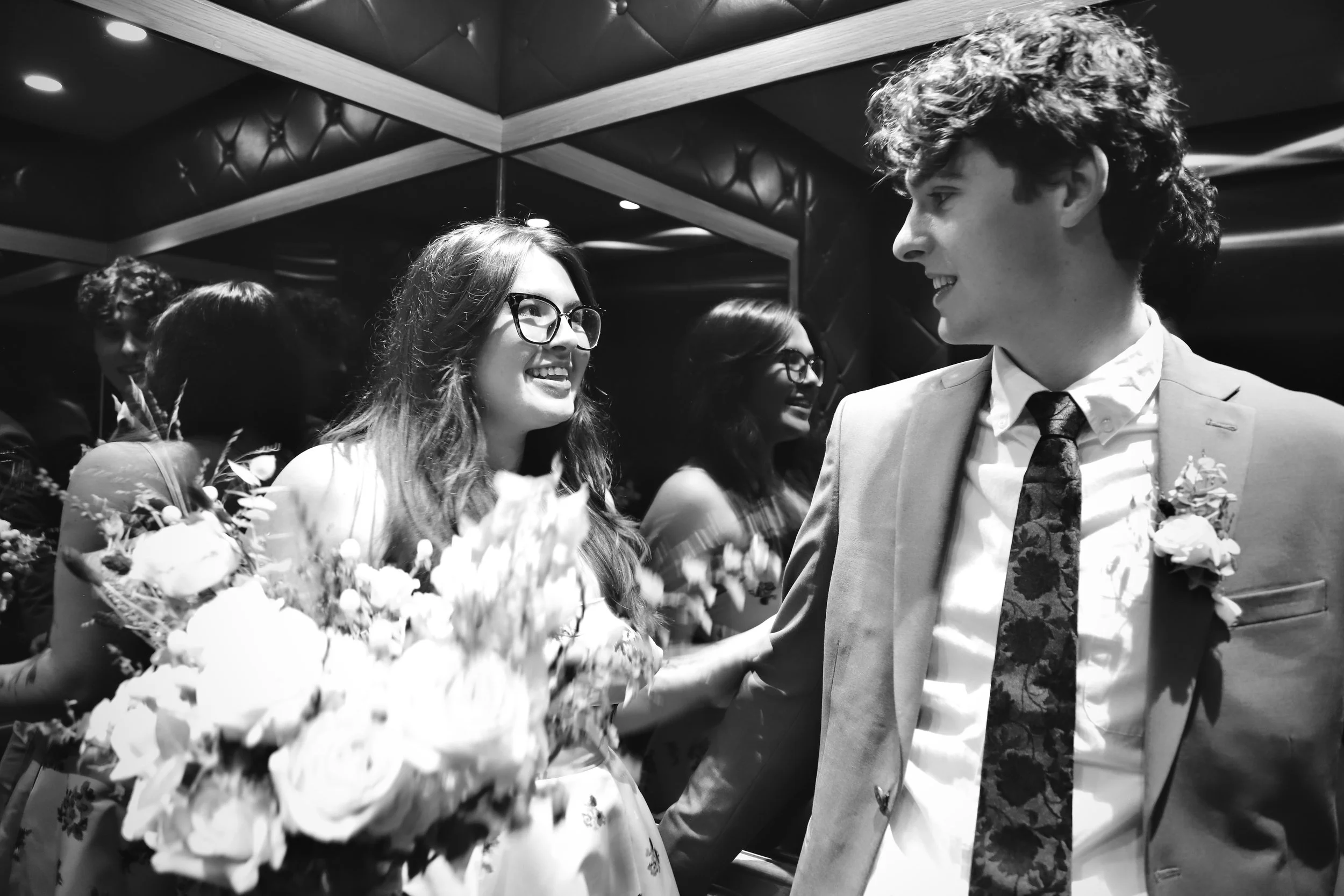 A black and white photo of a wedding reception with a bride holding a bouquet of flowers, smiling and looking at a groom in a suit. Several guests are in the background, smiling and enjoying the celebration.