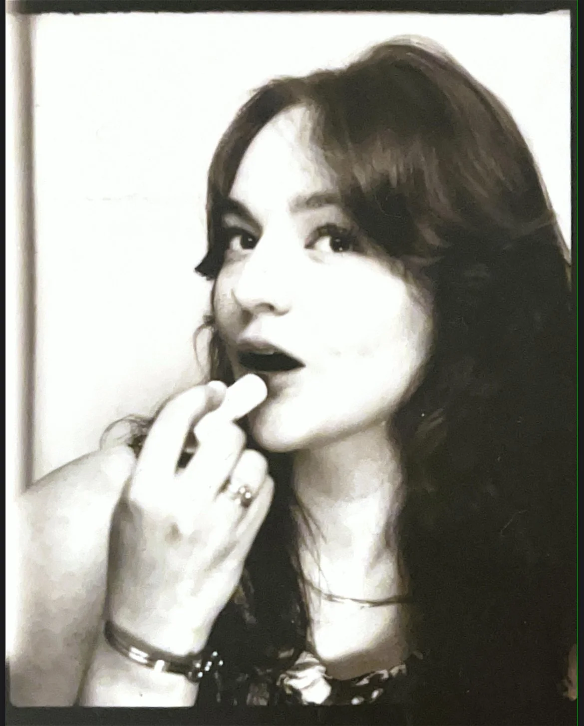 Black and white photo of a woman with long hair, wearing dark lipstick, touching her lips with her fingers, and looking slightly to the side.