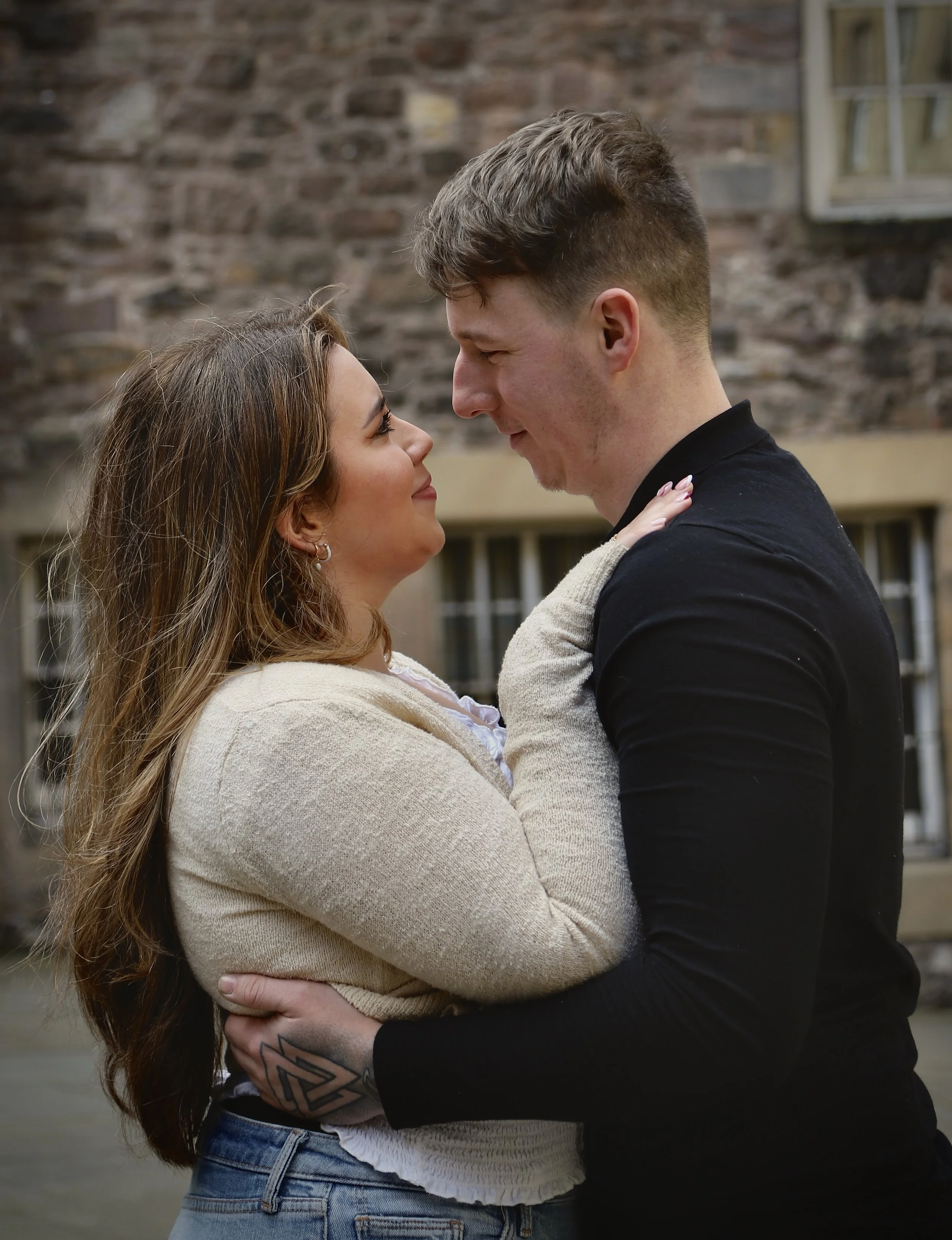 A couple standing close together, gazing into each other's eyes, with a brick building in the background.