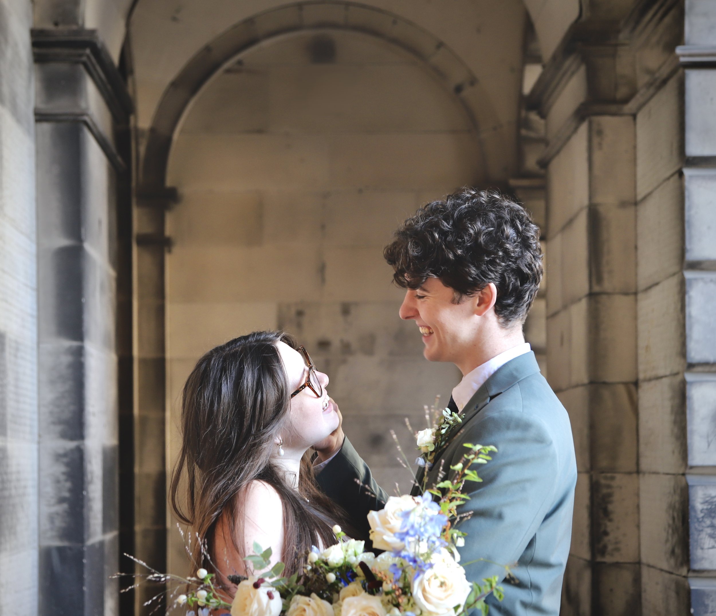 A smiling man and woman embrace inside a stone archway, with the woman holding a bouquet of flowers.