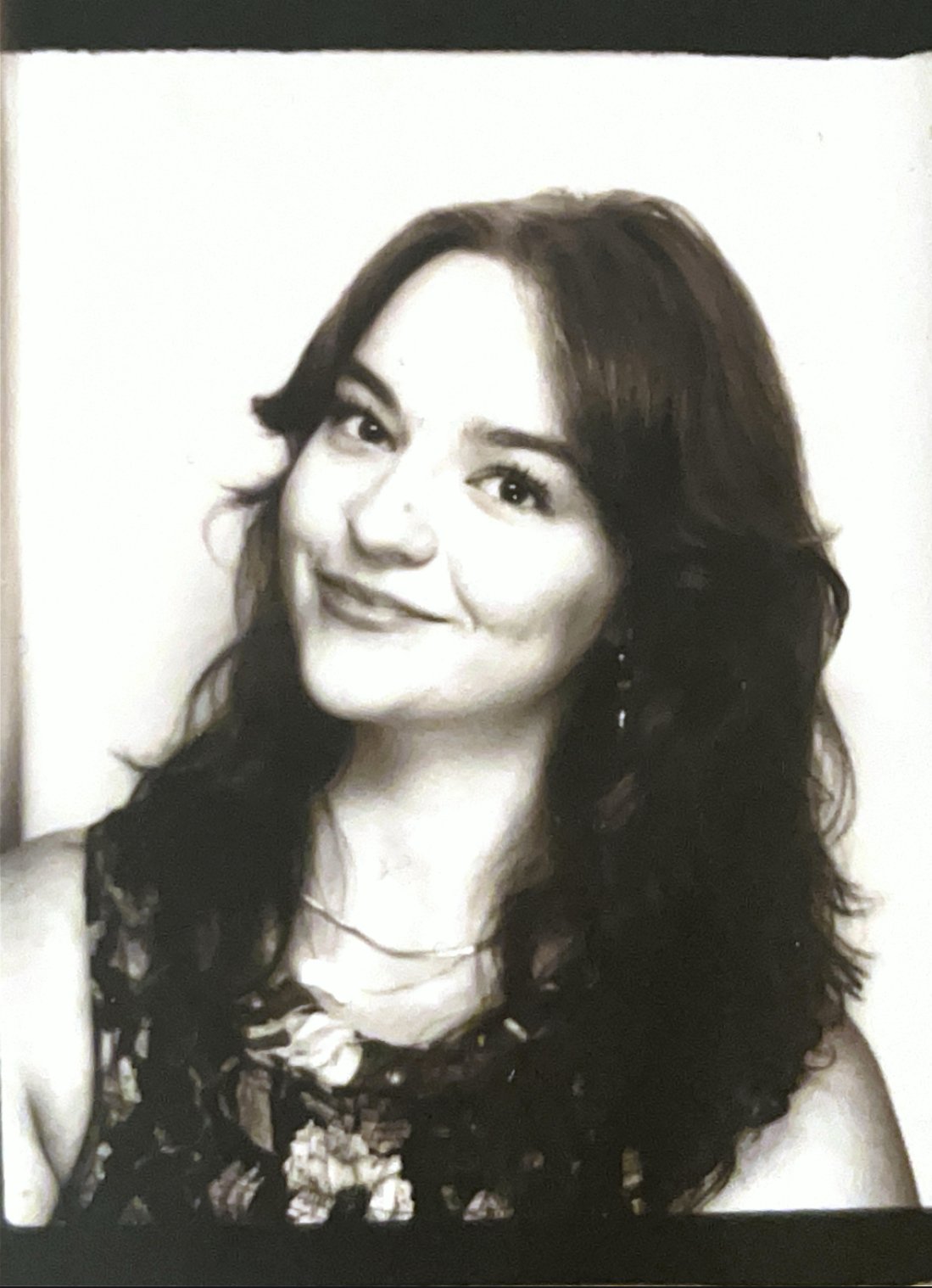 Black and white photograph of a woman with wavy dark hair, smiling, wearing earrings and a floral patterned top.