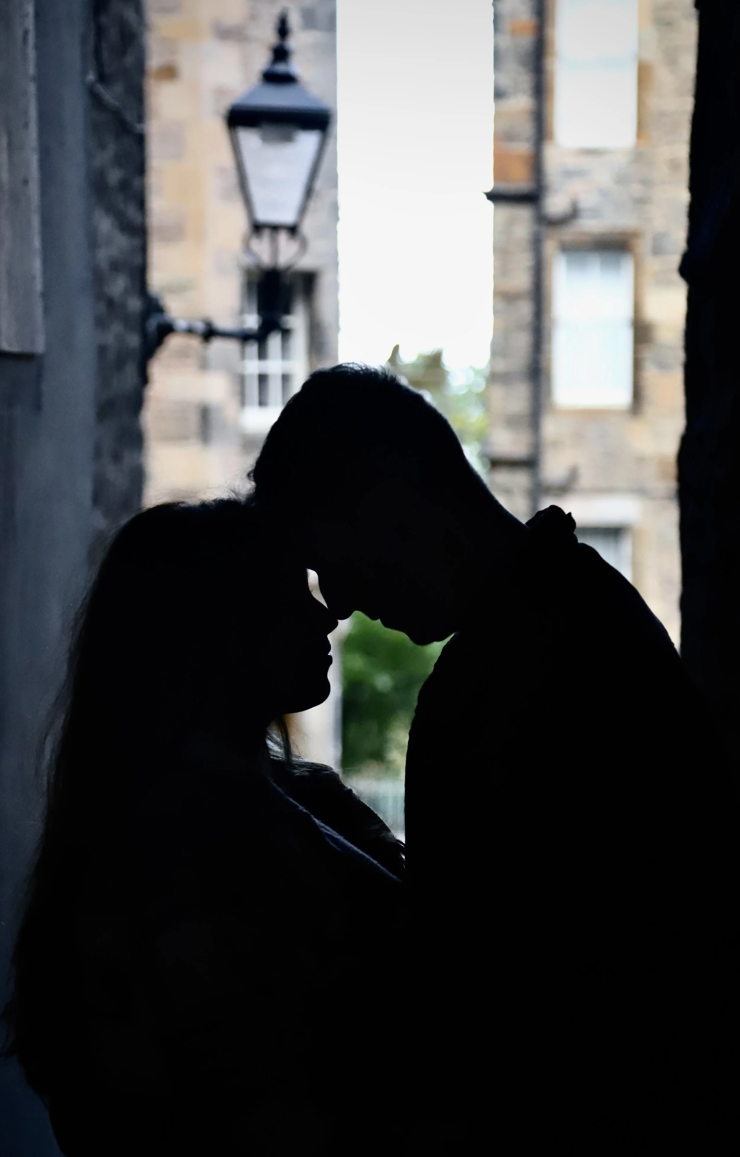 Silhouetted couple in a close, intimate pose, their foreheads touching, standing in a narrow alleyway with a brick building, a lamp post, and a window in the background.