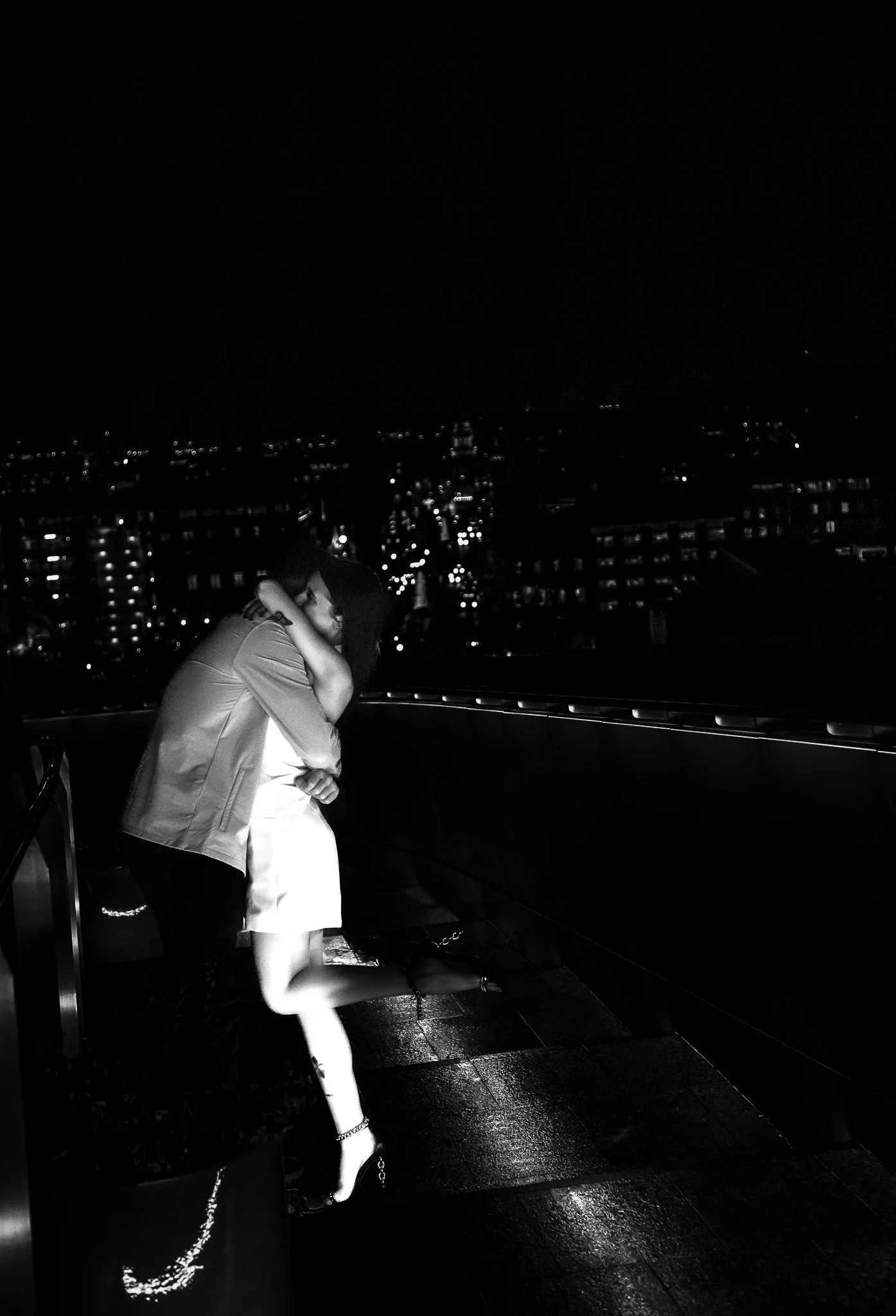 A couple of people embracing at night on a balcony overlooking a city skyline with lights, with one person standing on one leg while hugging another person.