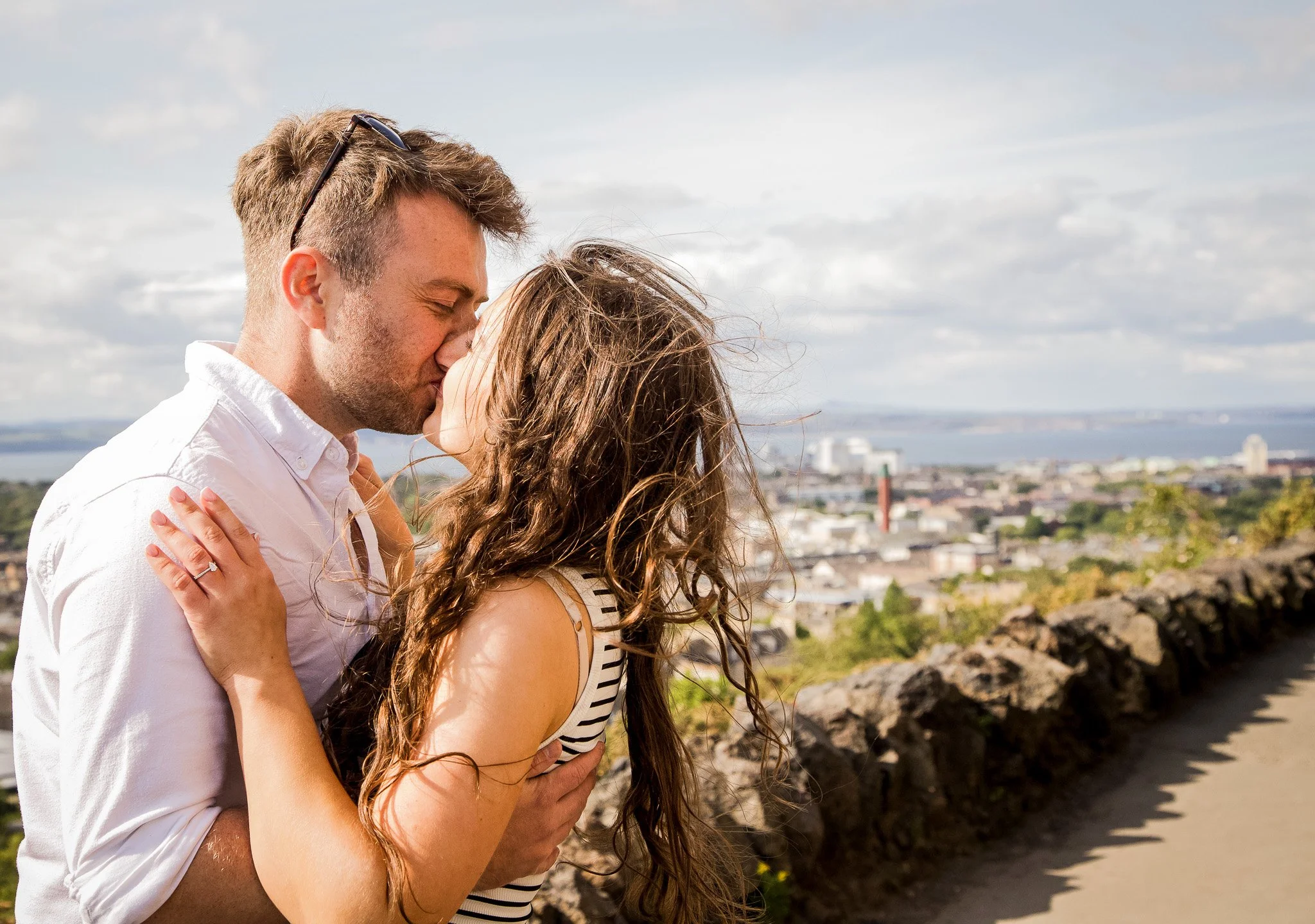 A couple standing outdoors on a sunny day, about to kiss, with a cityscape and water in the background.