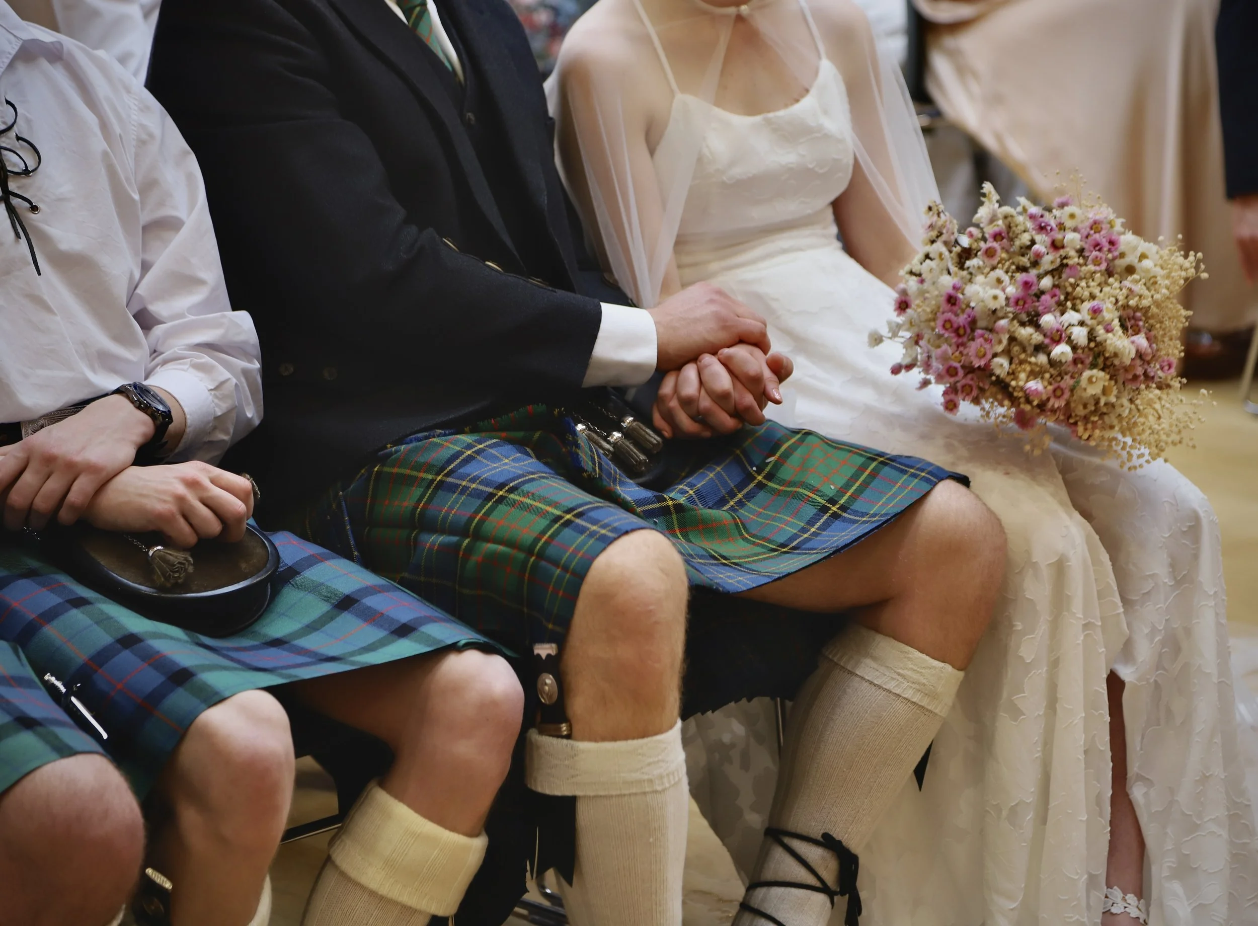 Wedding ceremony with a woman in a white wedding dress holding a bouquet of flowers, and men wearing traditional Scottish kilts with hands clasped.