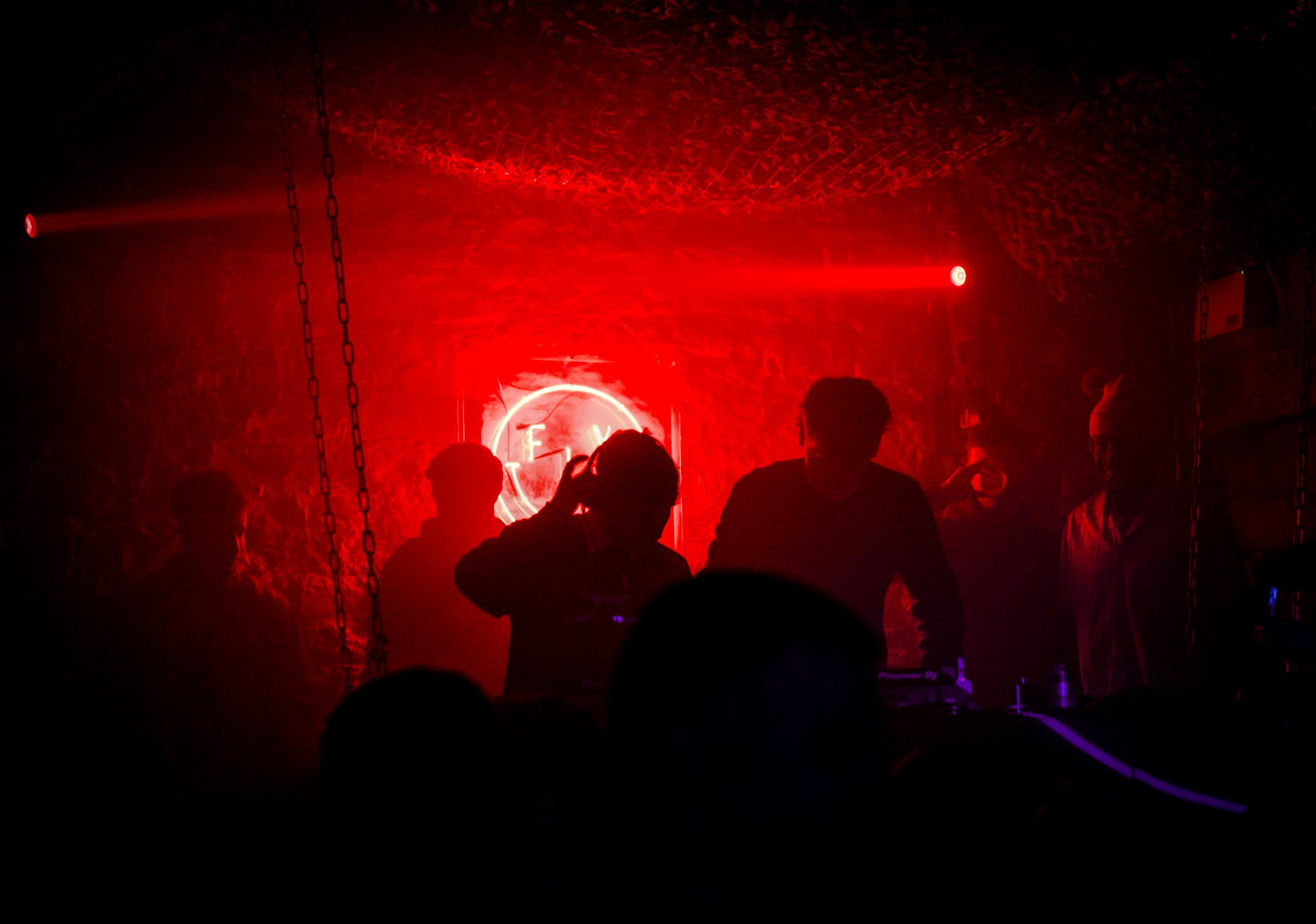 Silhouettes of people standing in a dimly lit room with red neon lights, some wearing headphones, with a neon sign on the brick wall behind them.