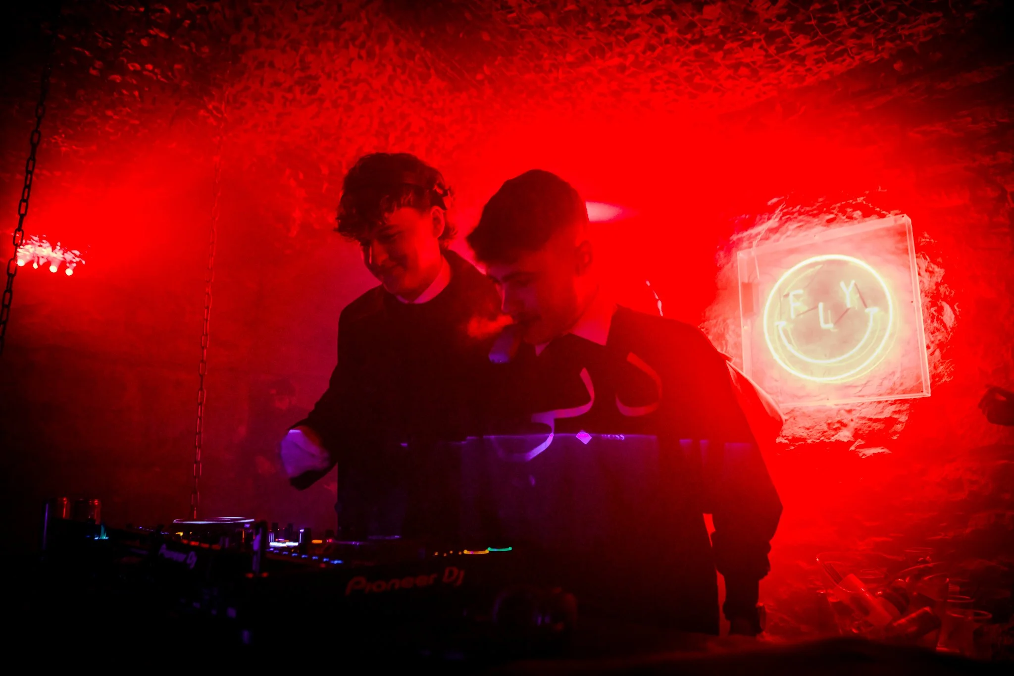 Two young men DJing in a dimly lit room with red lighting and a neon sign.