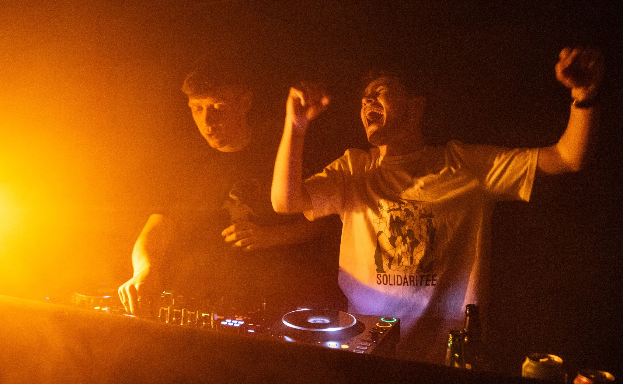 Two men DJing at a club or party, one wearing a T-shirt with the word 'Solidaritee', with orange lighting creating a vibrant atmosphere.