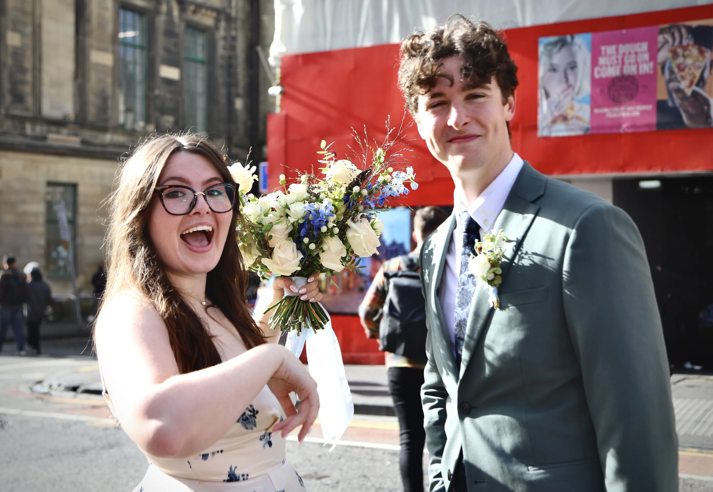 A celebrating couple on a city street, with the woman holding a bouquet of flowers and both smiling. The woman has long brown hair and glasses, wearing a cream-colored dress with blue floral patterns. The man has curly brown hair, dressed in a light gray suit with a boutonniere.