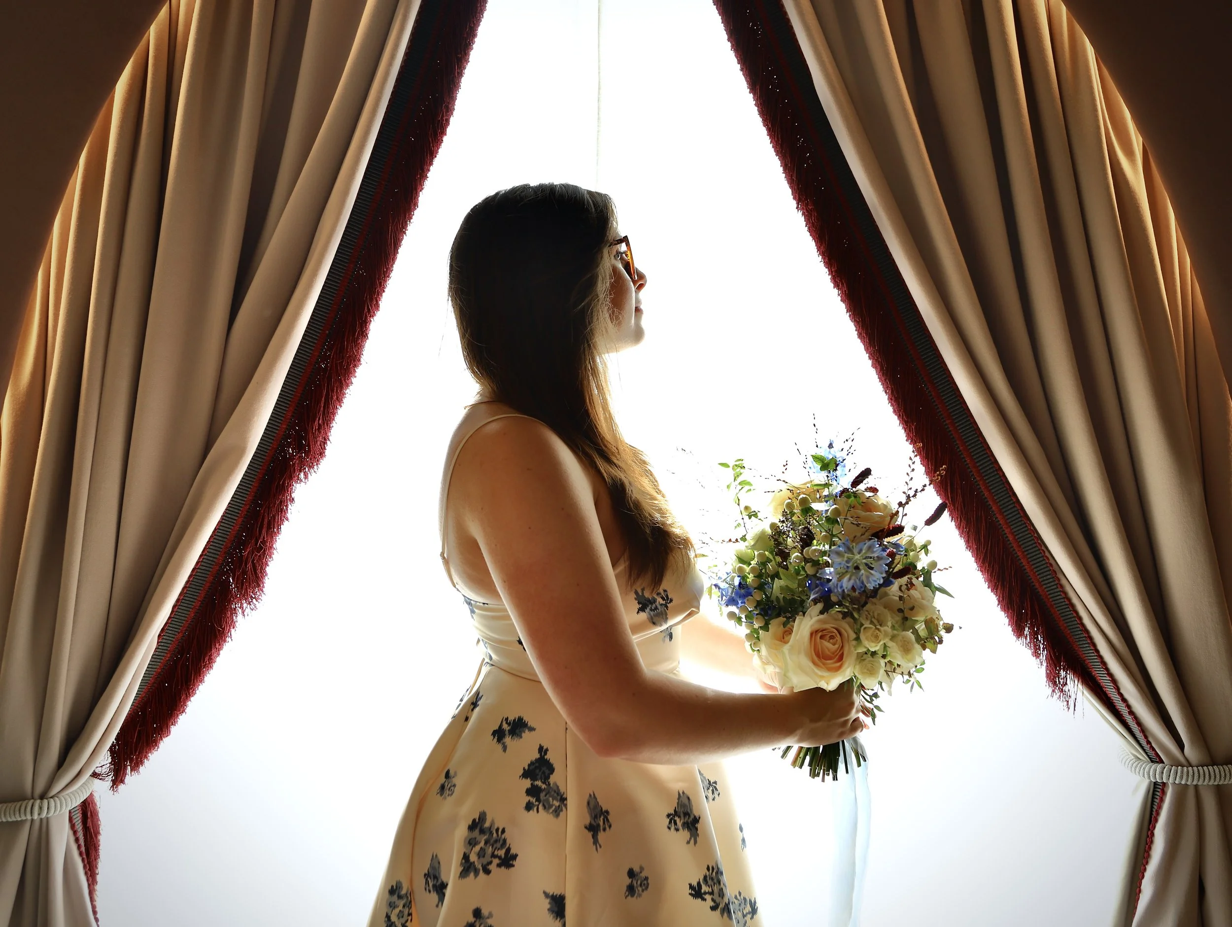 A woman standing near a window with red and beige curtains, holding a bouquet of flowers, looking outside.
