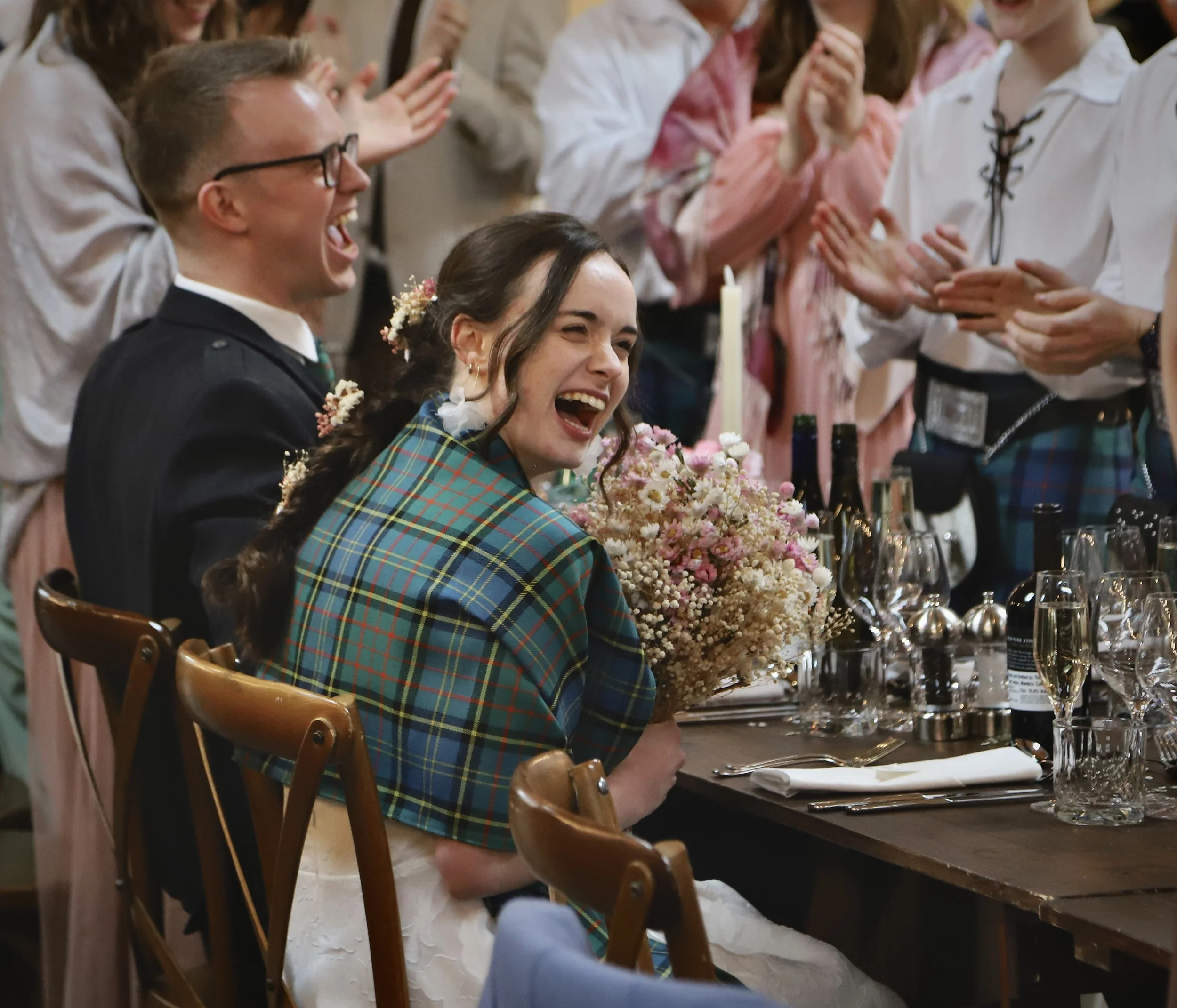 A group of people celebrating at a wedding reception, with a woman in a plaid dress holding a bouquet of flowers and laughing, surrounded by friends clapping and smiling.