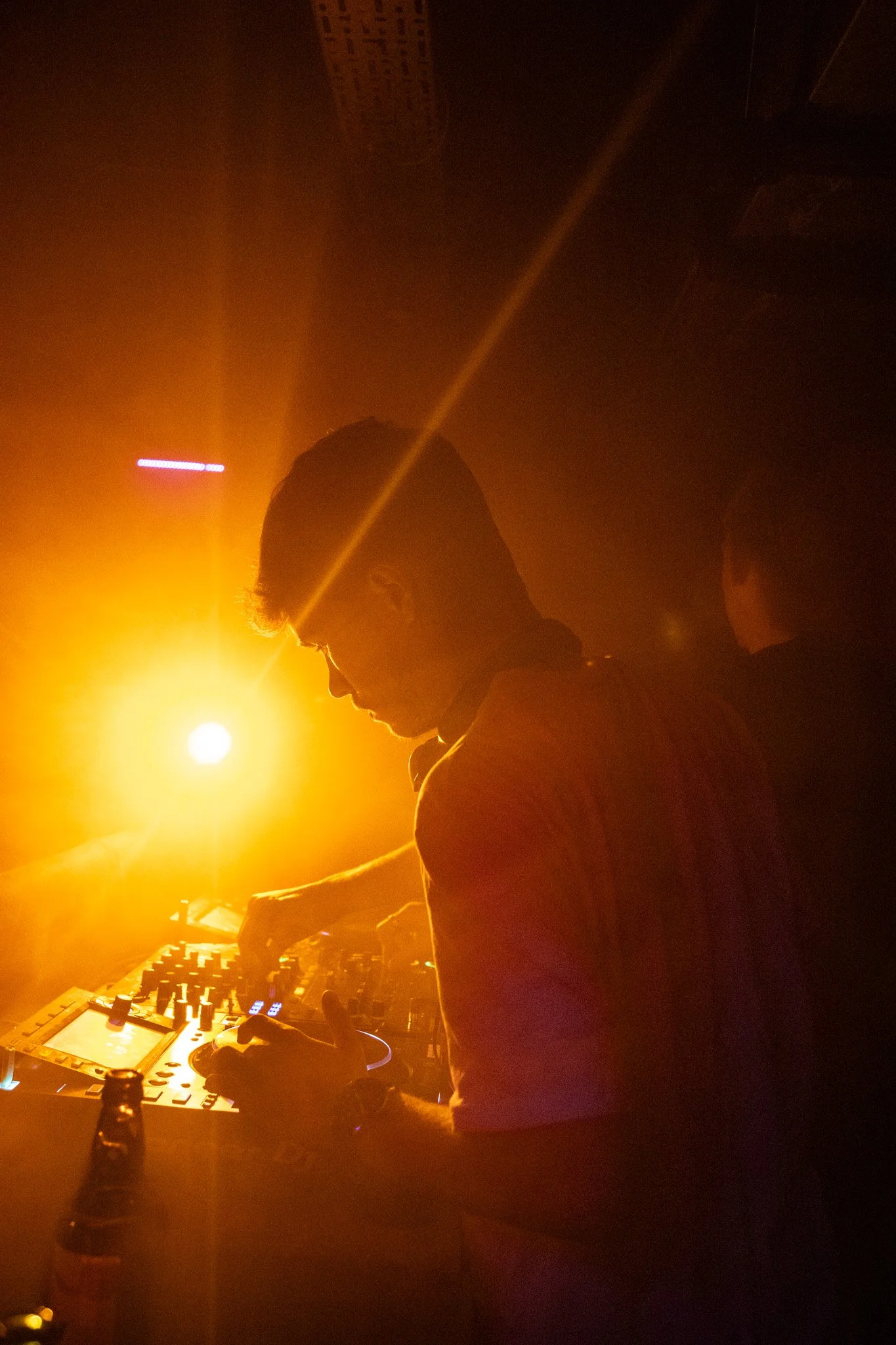 A person DJing in a dark club with orange and yellow lighting, using a mixer and turntables.