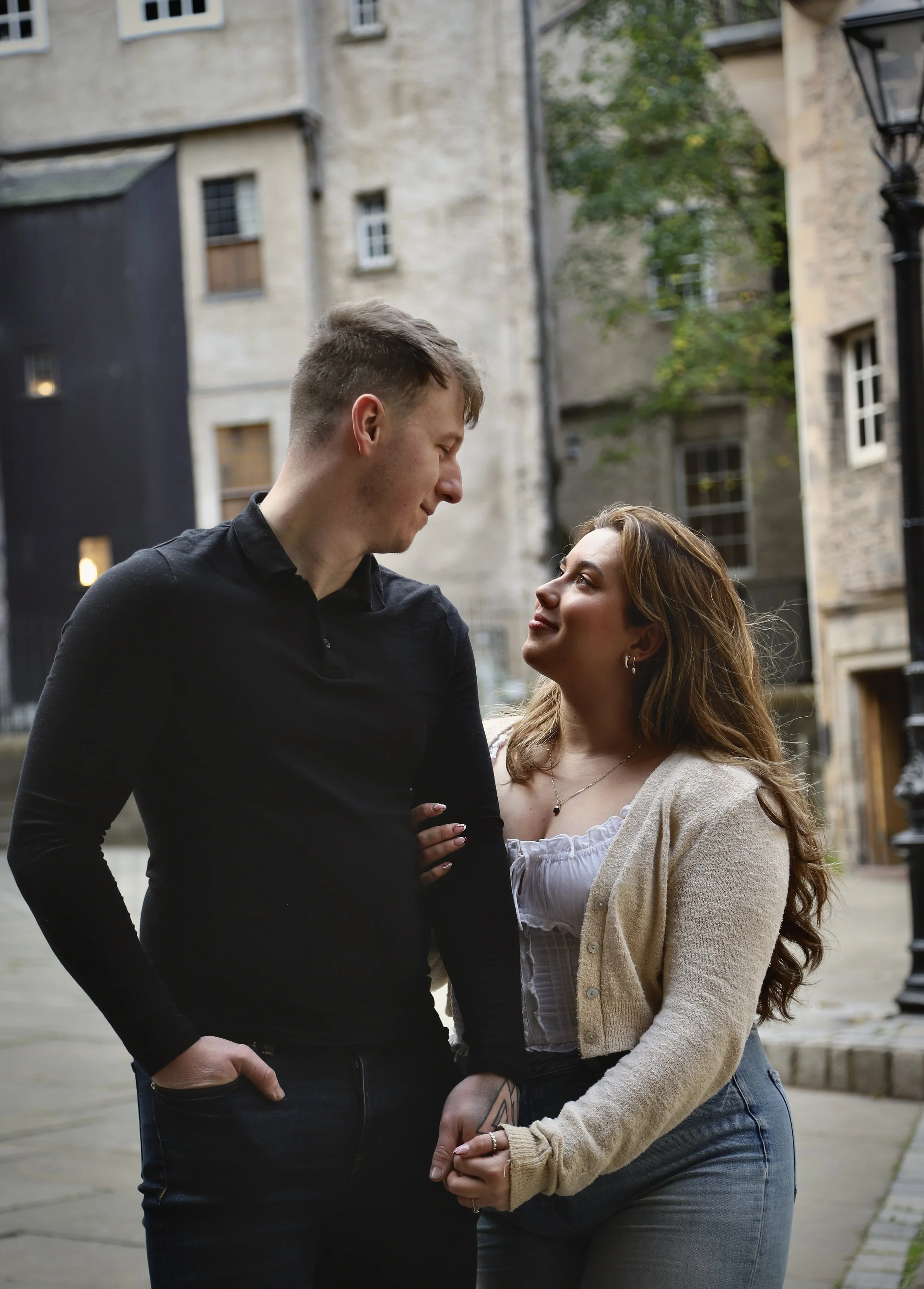 A man and woman standing close together outdoors, gazing into each other's eyes with the woman holding the man's hand, in an urban setting with old stone buildings and a lamppost in the background.
