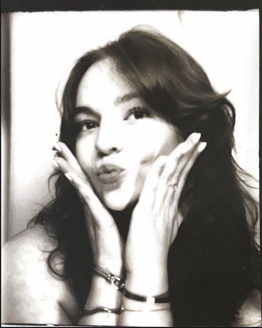 Black and white portrait of a woman with wavy hair, pursing her lips, and clasping her face with both hands.