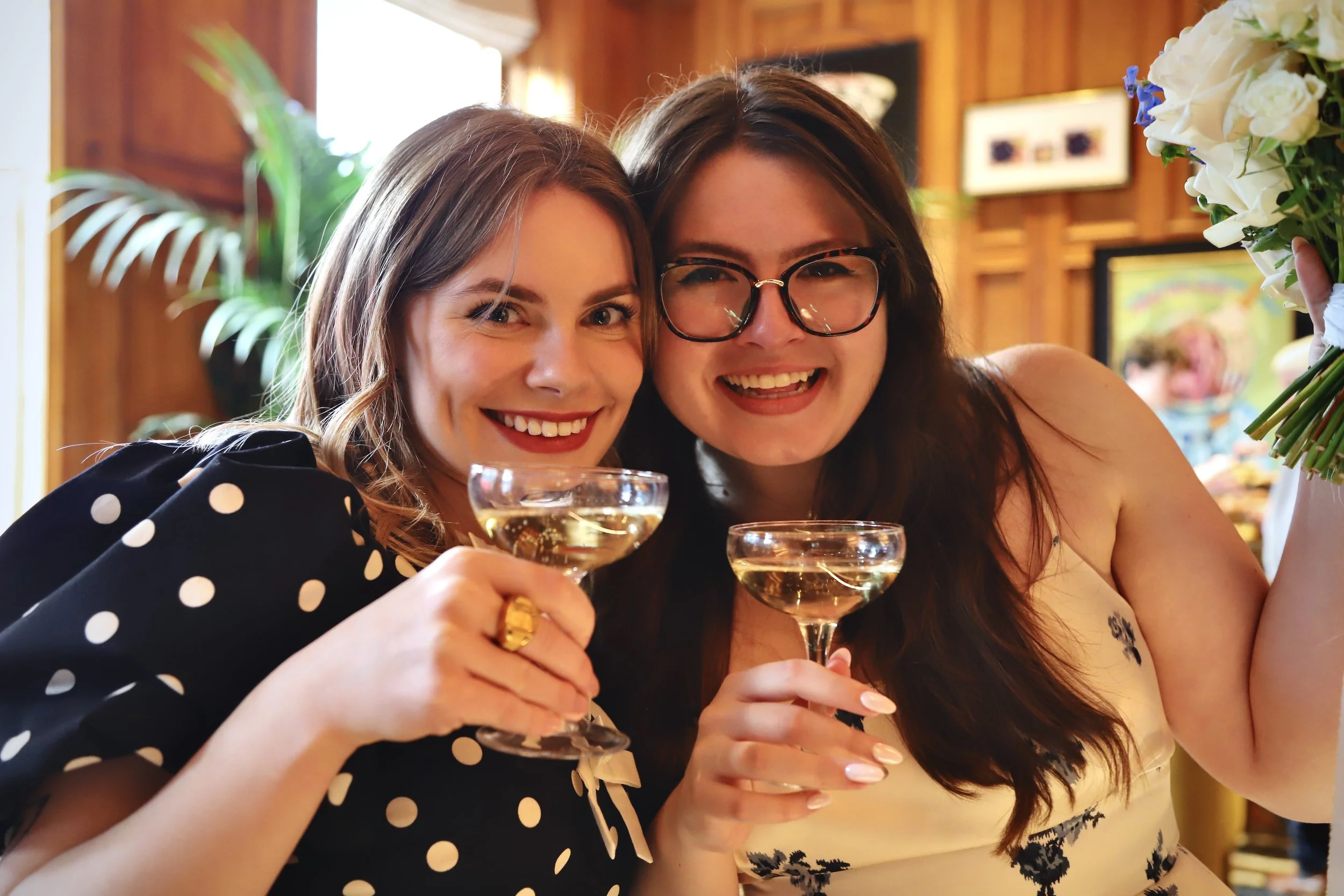 Two women smiling and holding champagne glasses at a celebration or party in a warmly lit room with wooden decor.