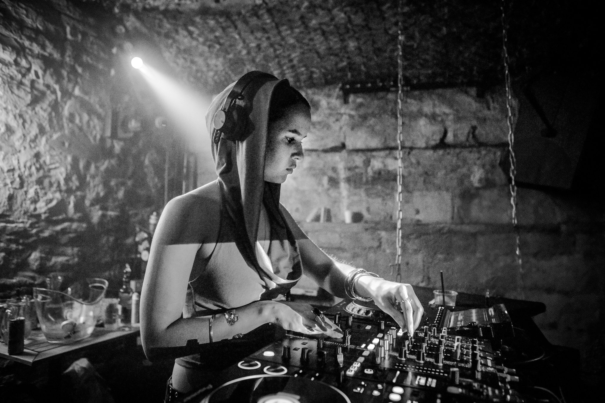 A woman DJ wearing headphones, working on a mixer, in a dimly lit, rocky underground setting.