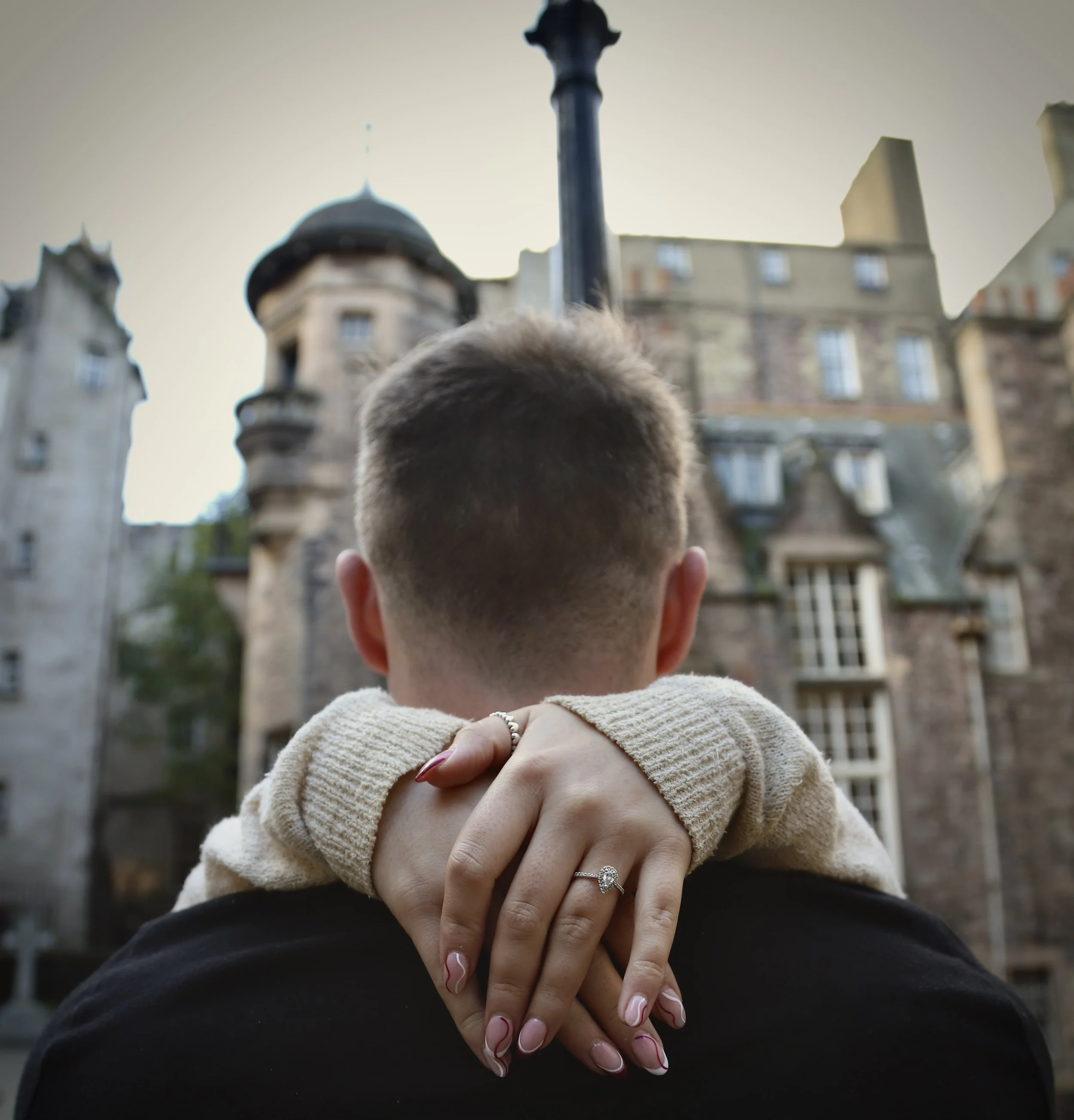 A woman’s hand with an engagement ring is resting on a man’s shoulder, facing away from the camera, with a historic building in the background.