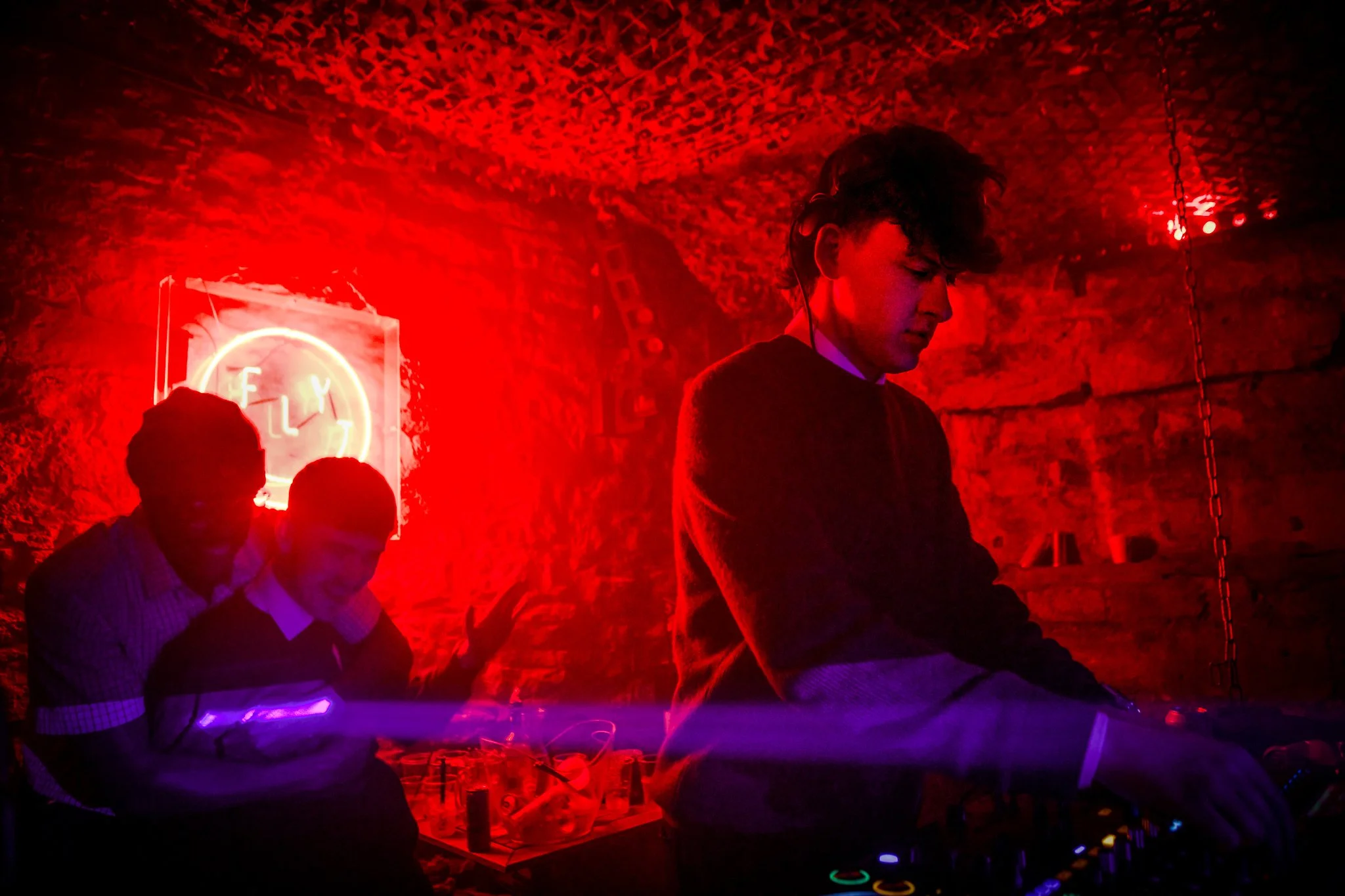 A DJ performs at a nightclub with red lighting, a neon sign behind him, and two people sitting nearby.