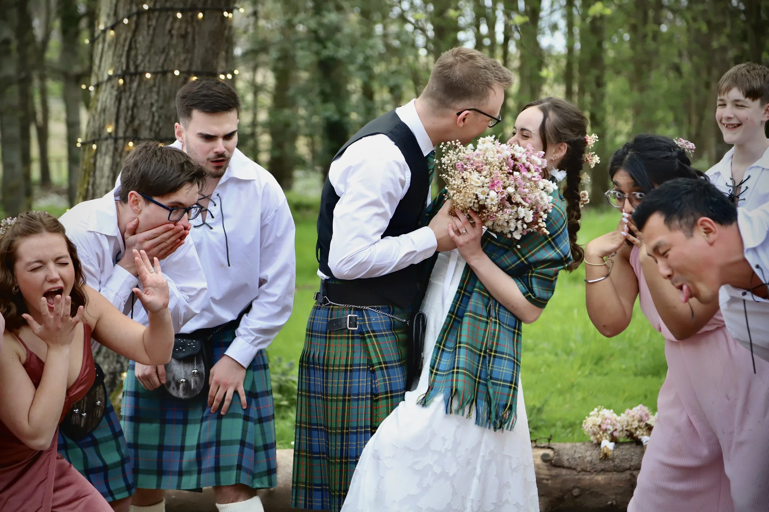 A wedding scene in the woods with a bride and groom holding a bouquet of flowers surrounded by friends reacting with surprise and excitement.