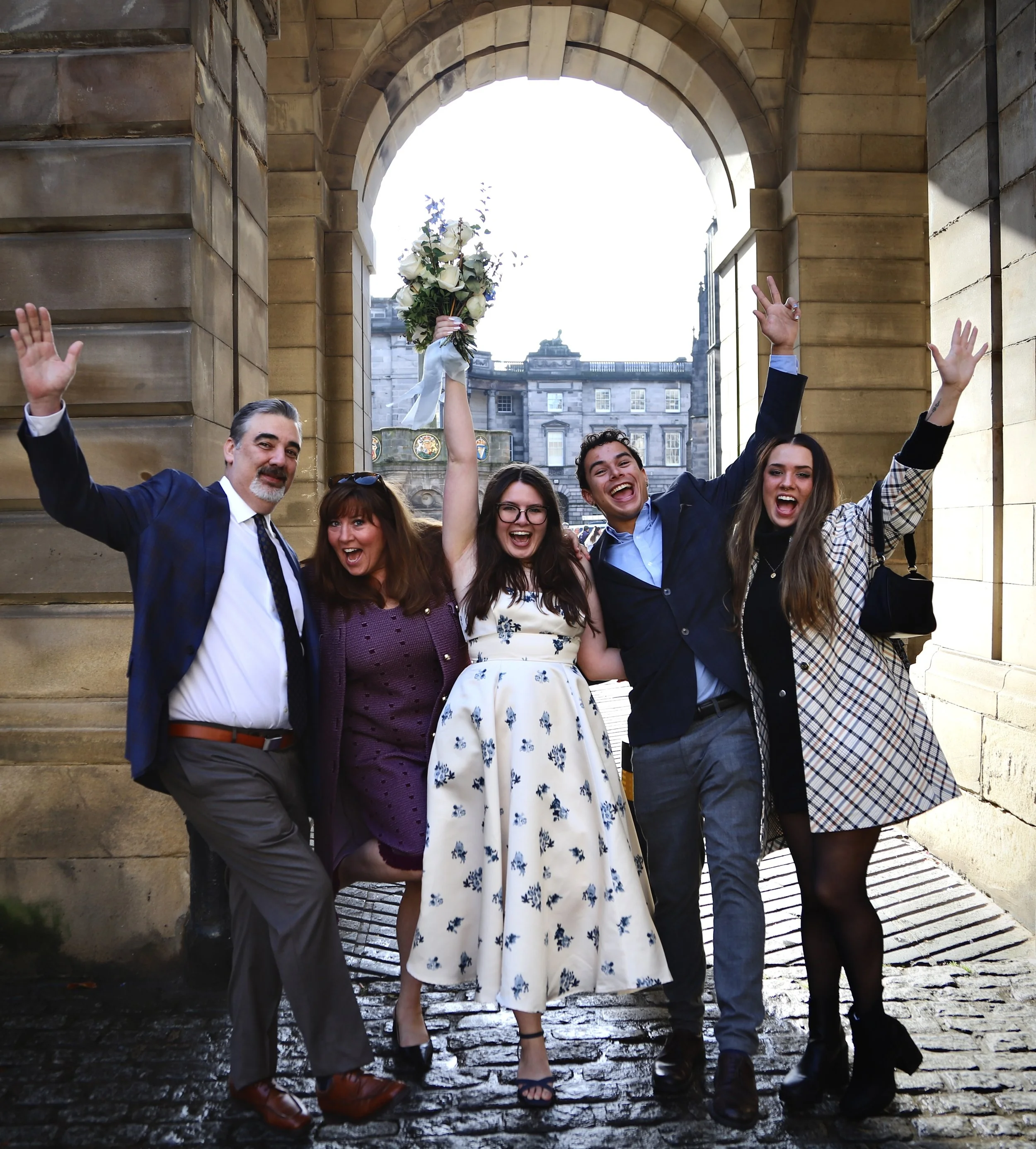 A group of five friends celebrating outdoors under an archway, with one woman holding a bouquet and all smiling and raising their hands.