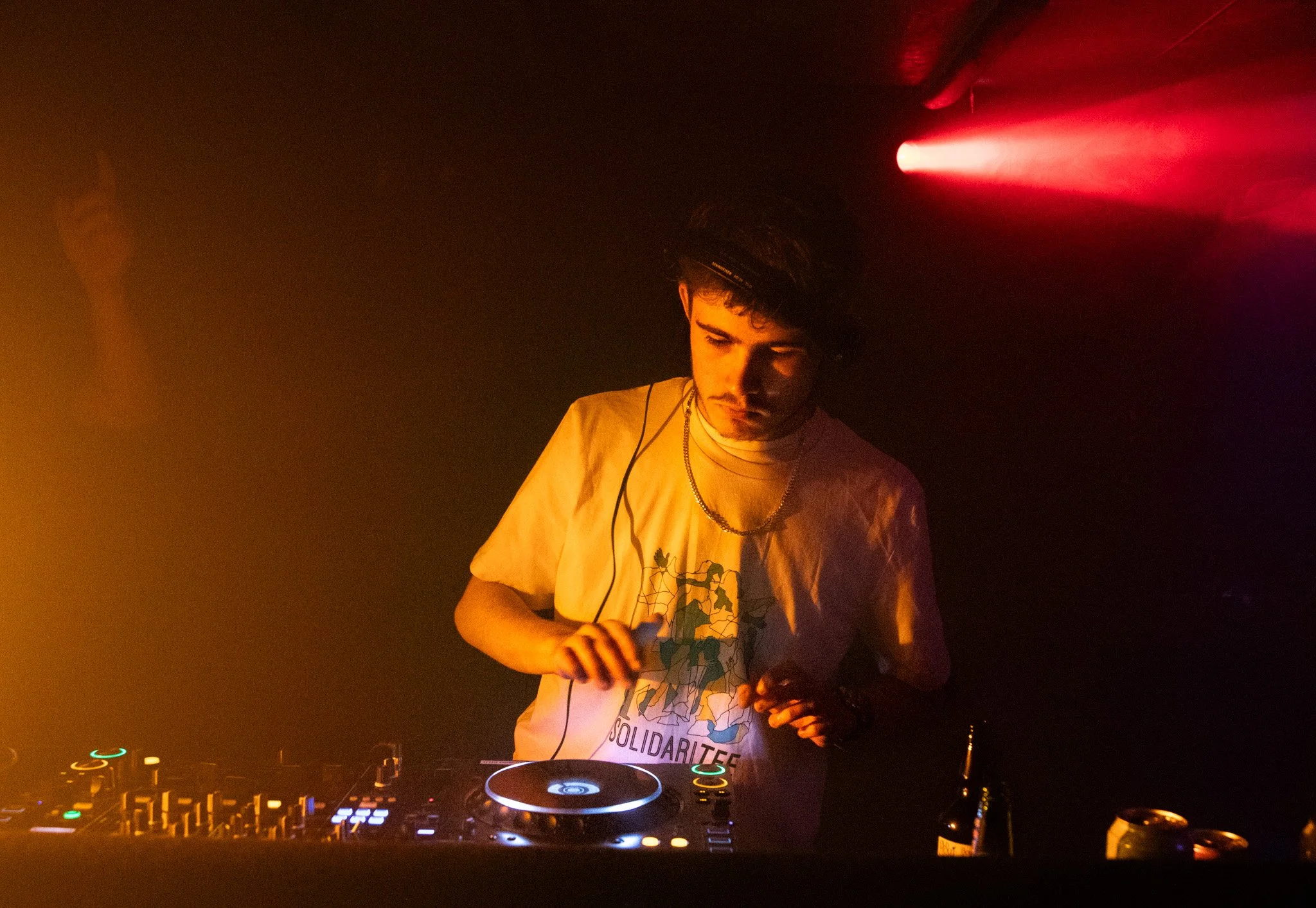 A DJ with dark hair and a mustache wearing a beige T-shirt with a graphic design and a gold chain, standing behind DJ equipment in a dimly lit nightclub with red and yellow lighting.