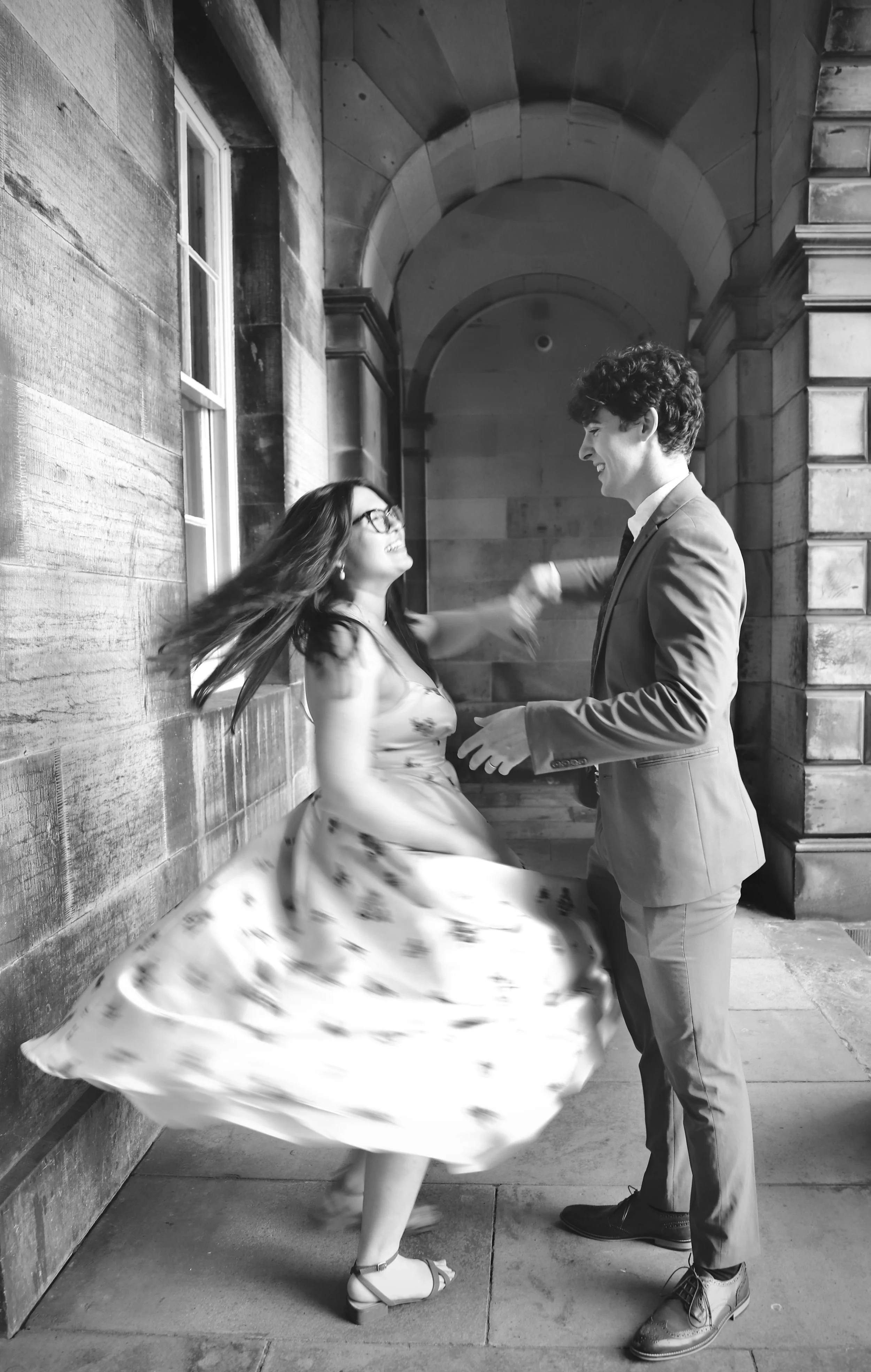 A black and white photo of a woman spinning around in a floral dress and heels, and a man in a suit smiling at her as they dance or share a joyful moment outside near a stone building.