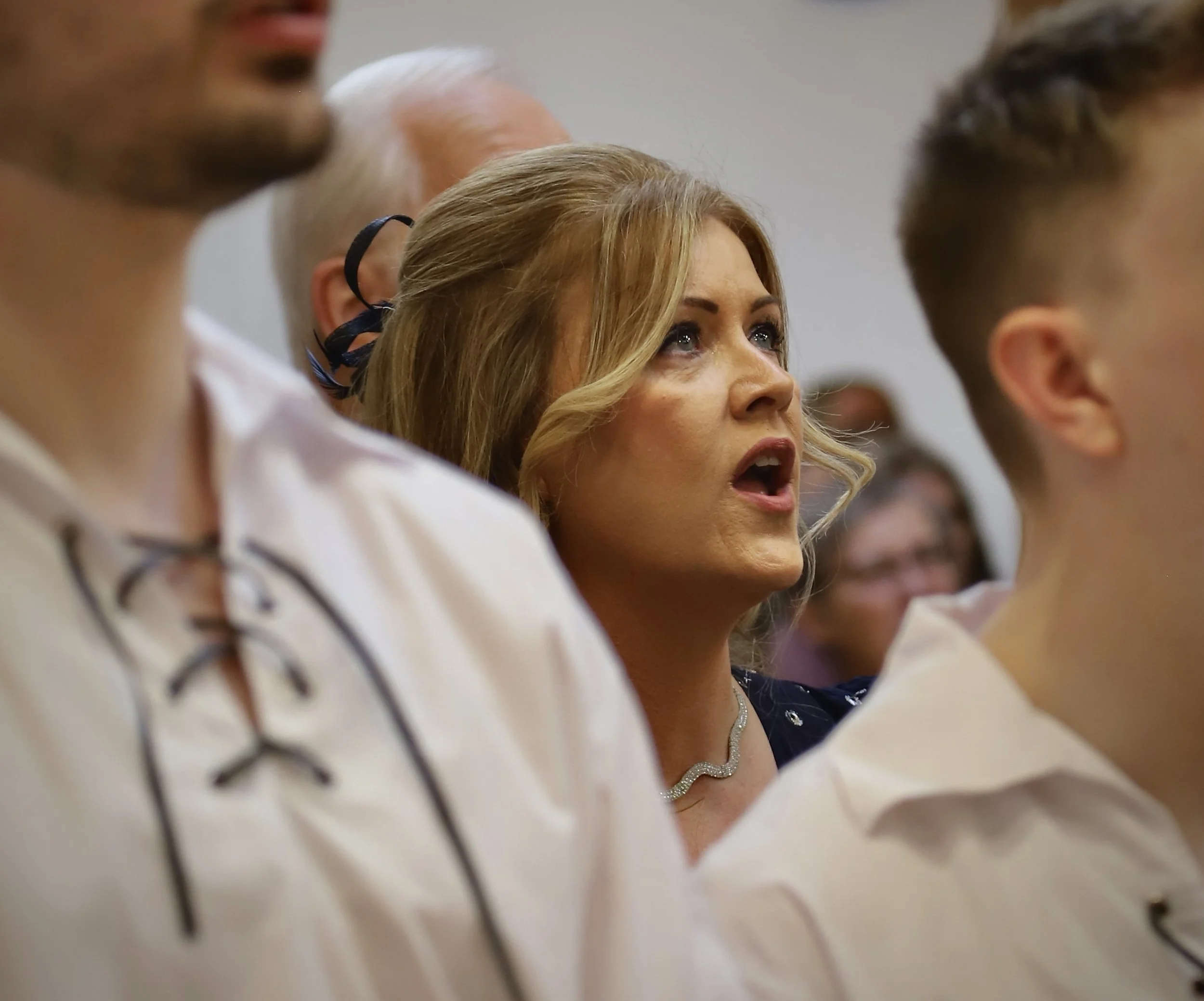 A woman with light brown hair, wearing a diamond necklace, looks surprised or emotional during an event, seated among other people.