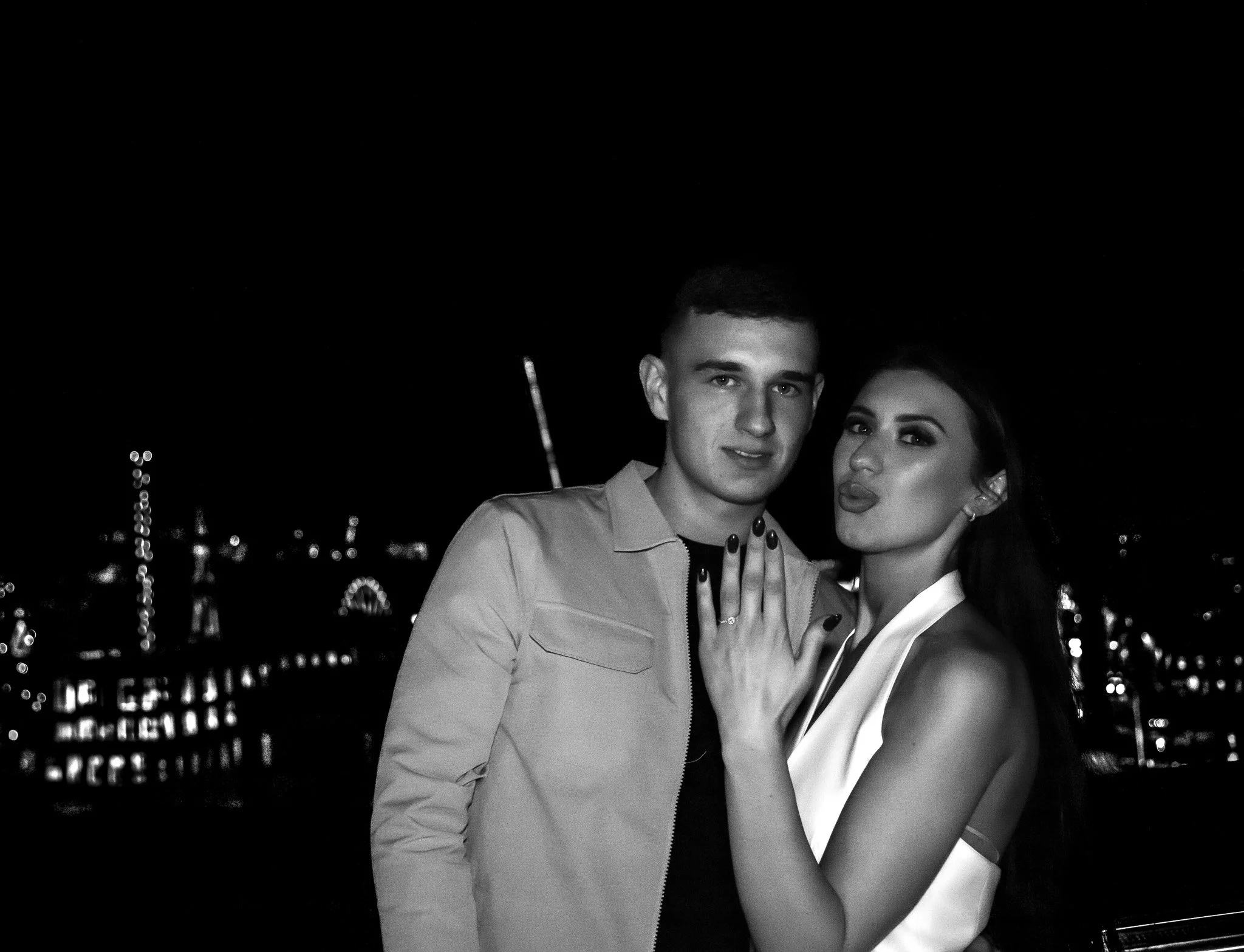 A black and white photo of a young man and woman standing close together at night with city lights and ferris wheel in the background. The woman shows off her ring on her left hand.