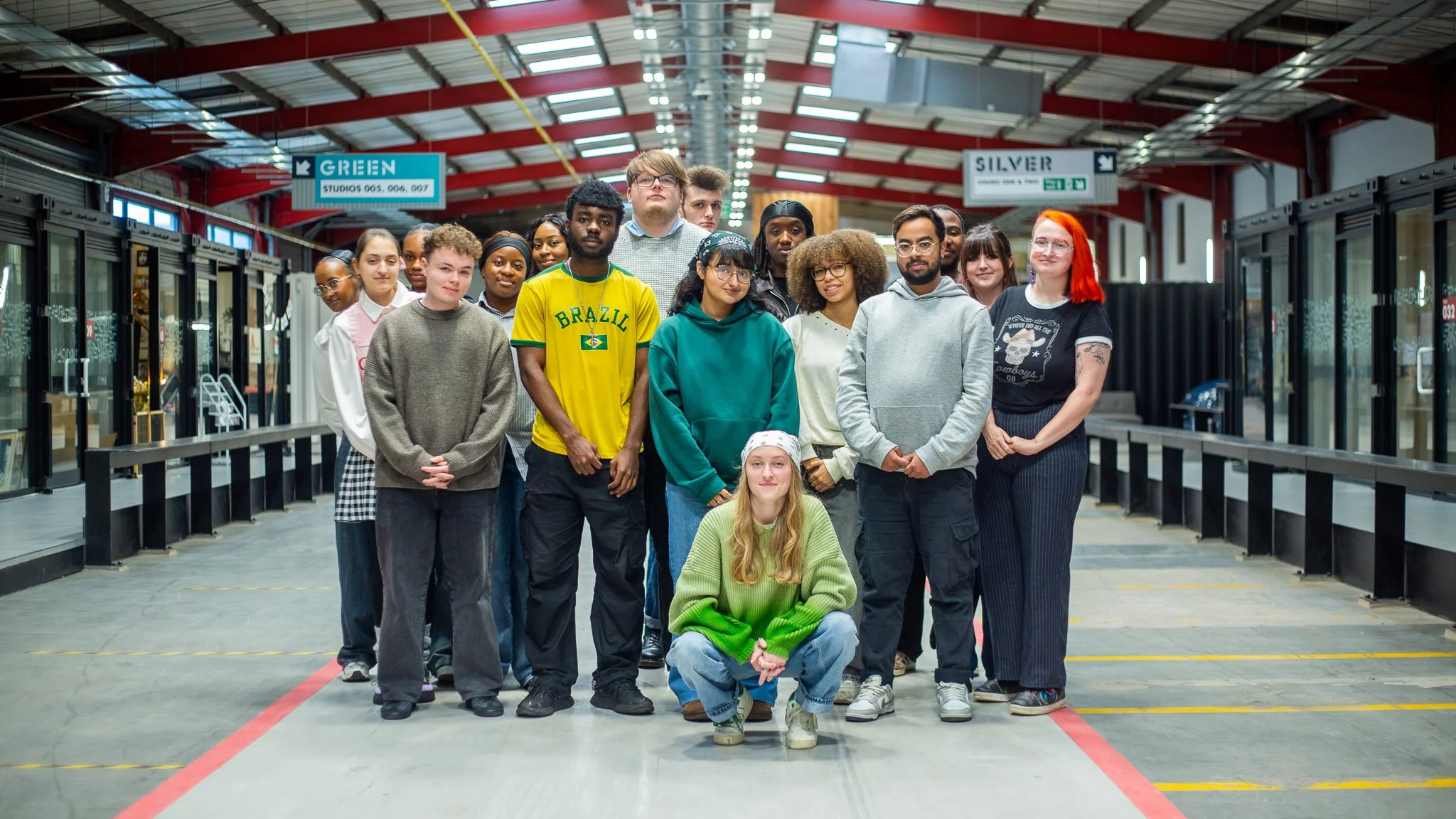Group of young diverse people standing in an industrial-style indoor space with a sign overhead indicating different studio rooms, posing for a photo.