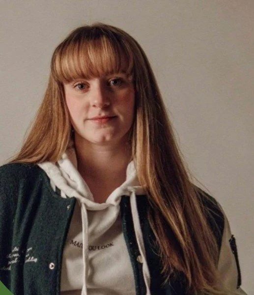 A young woman with long brown hair and bangs, wearing a white hoodie and a dark jacket, standing against a plain wall.