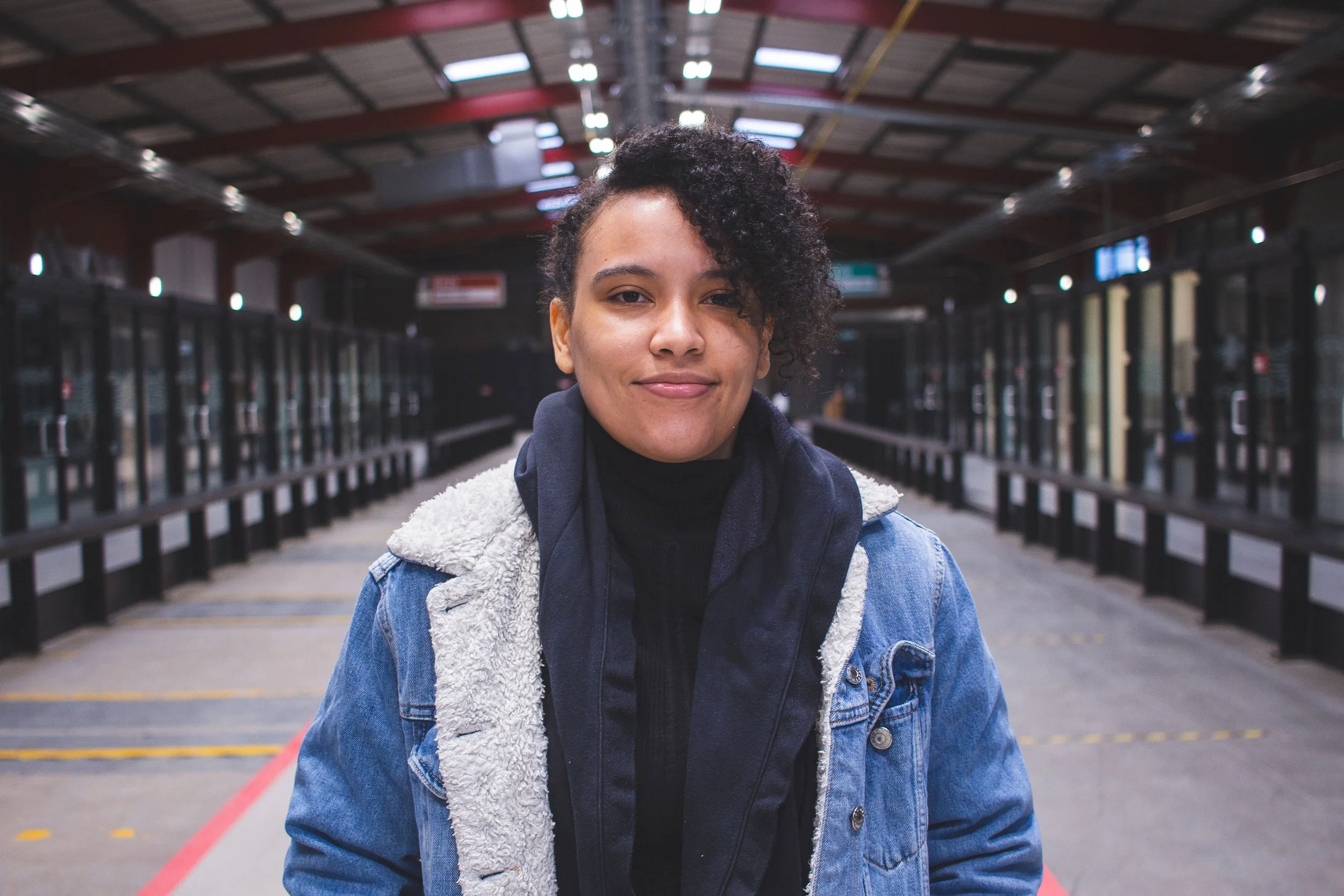 A woman with dark curly hair wears a black turtleneck and a denim jacket with a sherpa collar, standing inside a covered train or bus station with glass walls and a high arched ceiling.