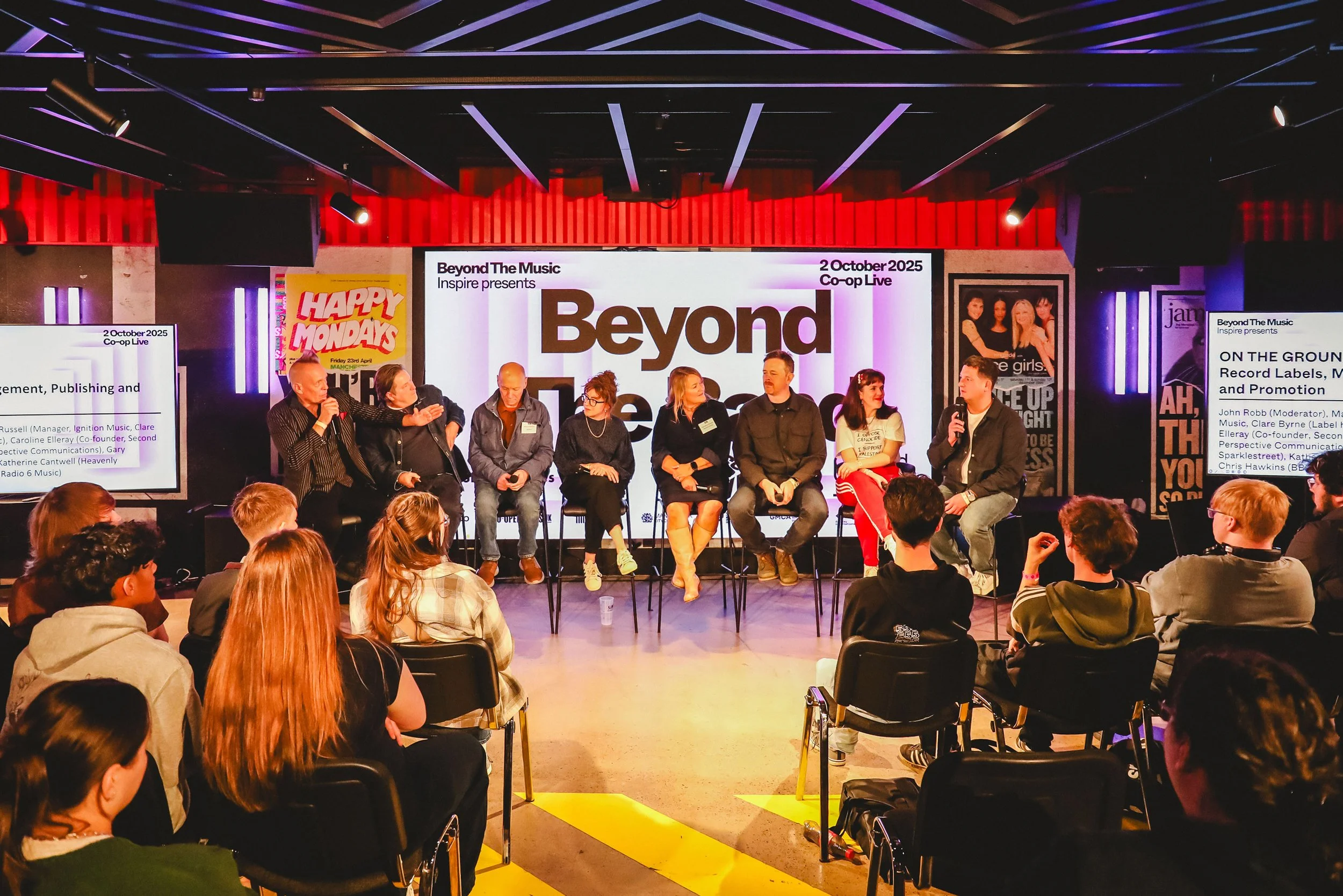Panel discussion at a conference with nine people on stage, four women and five men, seated on stools with microphones, audience listening, large screen behind displaying 'Beyond The Music' and other text, posters on walls, dim lighting with spotlights