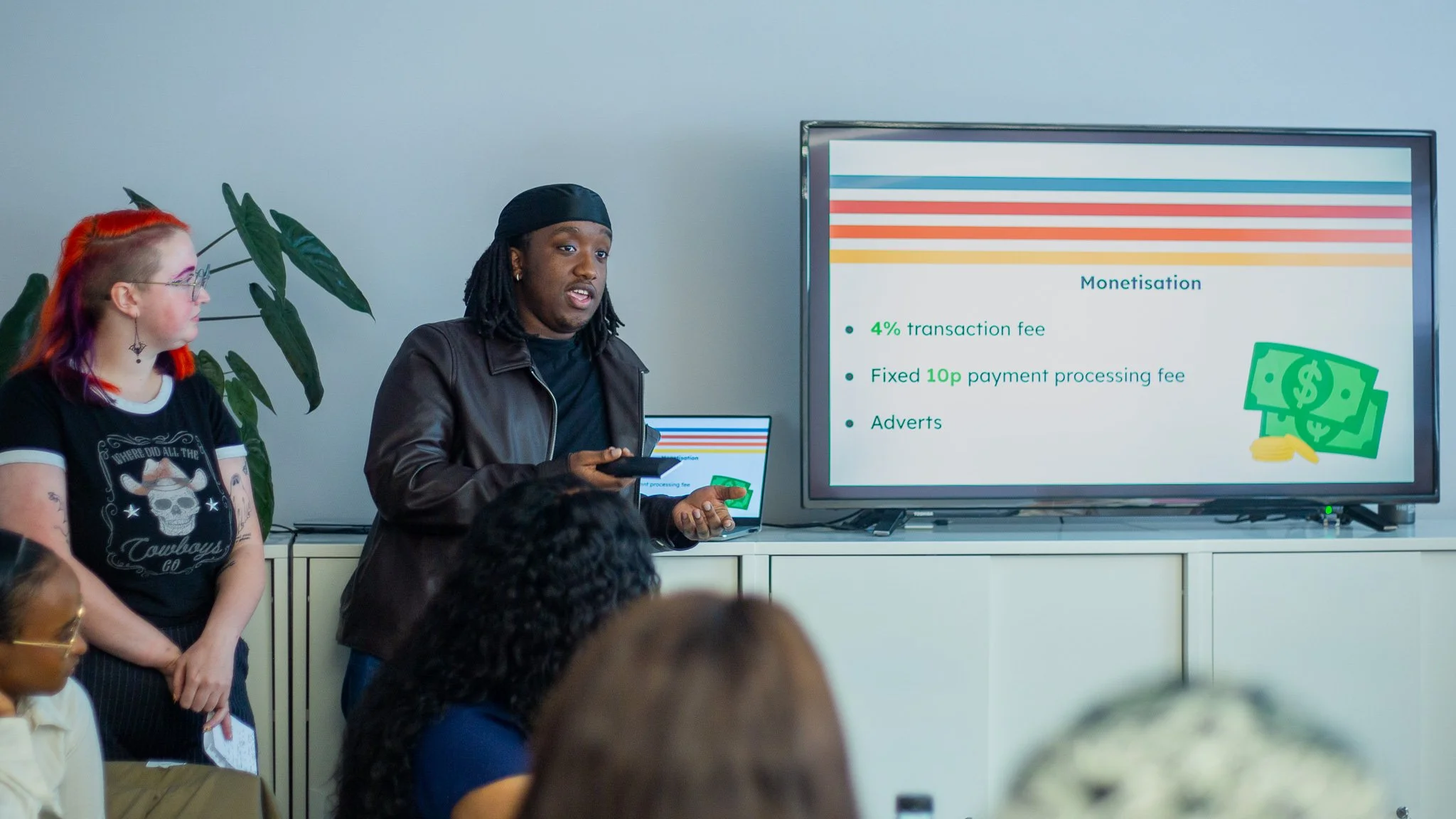 A man in a brown leather jacket and black head covering is giving a presentation about monetisation to a group of people, with a large screen displaying bullet points and an illustration of dollar bills.