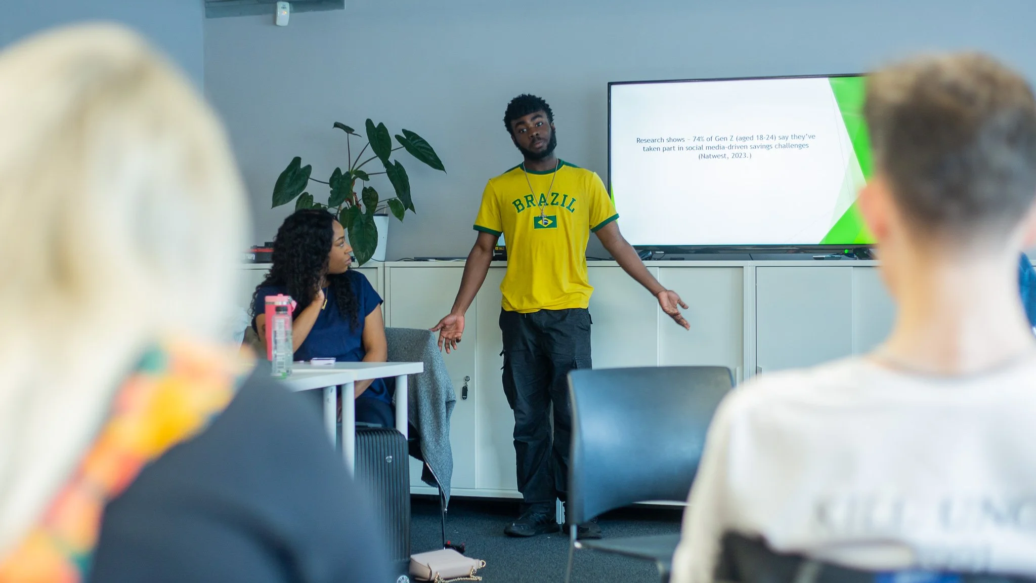 A man wearing a Brazil football jersey is giving a presentation in a classroom or conference room while standing in front of a TV screen. One woman is seated at a table and looking at him, and several other people are facing him, out of focus.