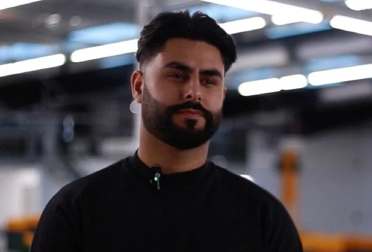 A man with dark hair and a beard wearing a black shirt in an indoor setting with industrial lighting.