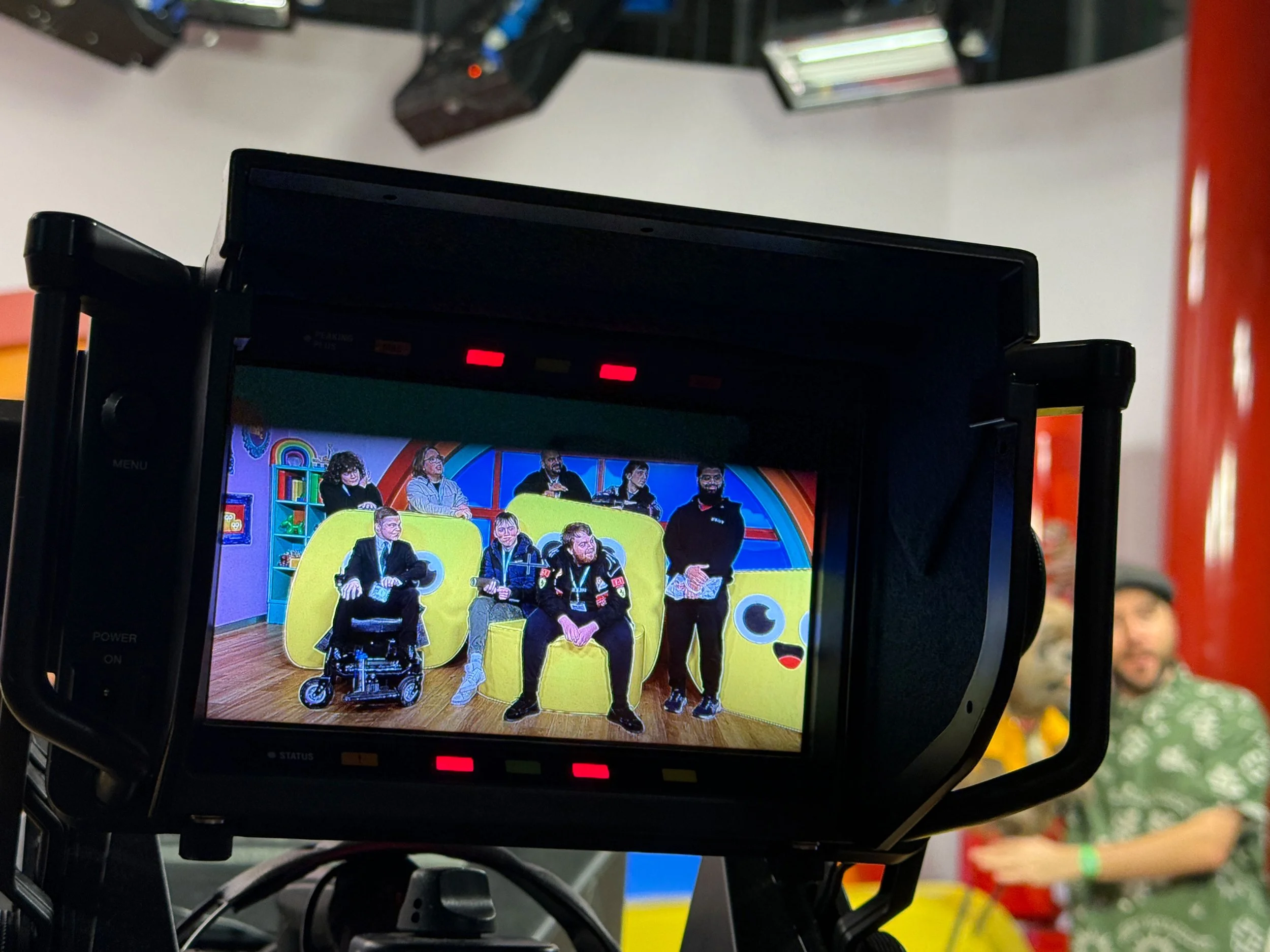 A TV camera captures a group of people on a colorful TV show set with various props and decorations, including a large yellow character and a red background.
