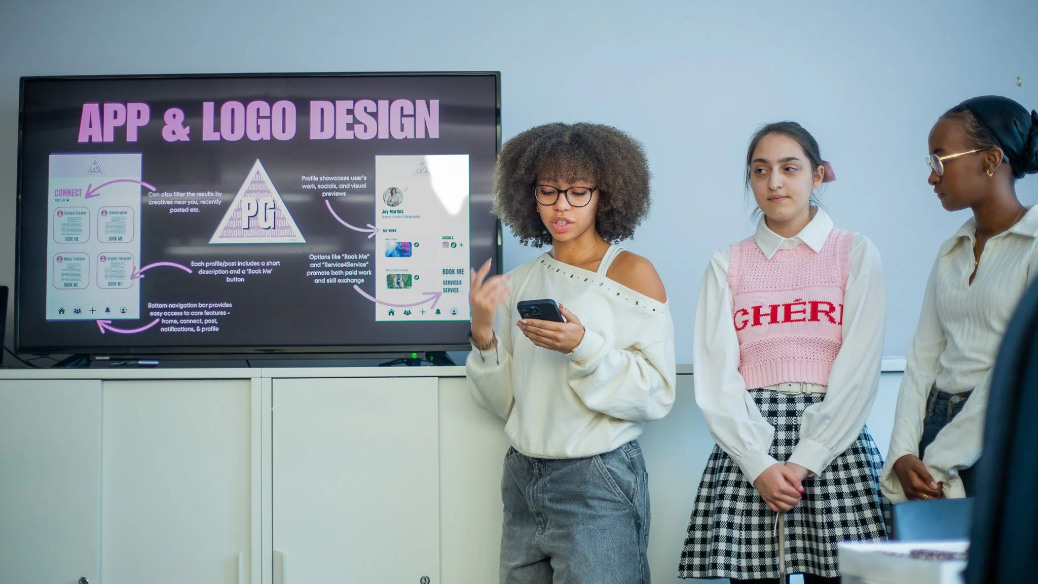 Three women standing in front of a presentation screen discussing app and logo design, with one woman speaking and holding a smartphone.