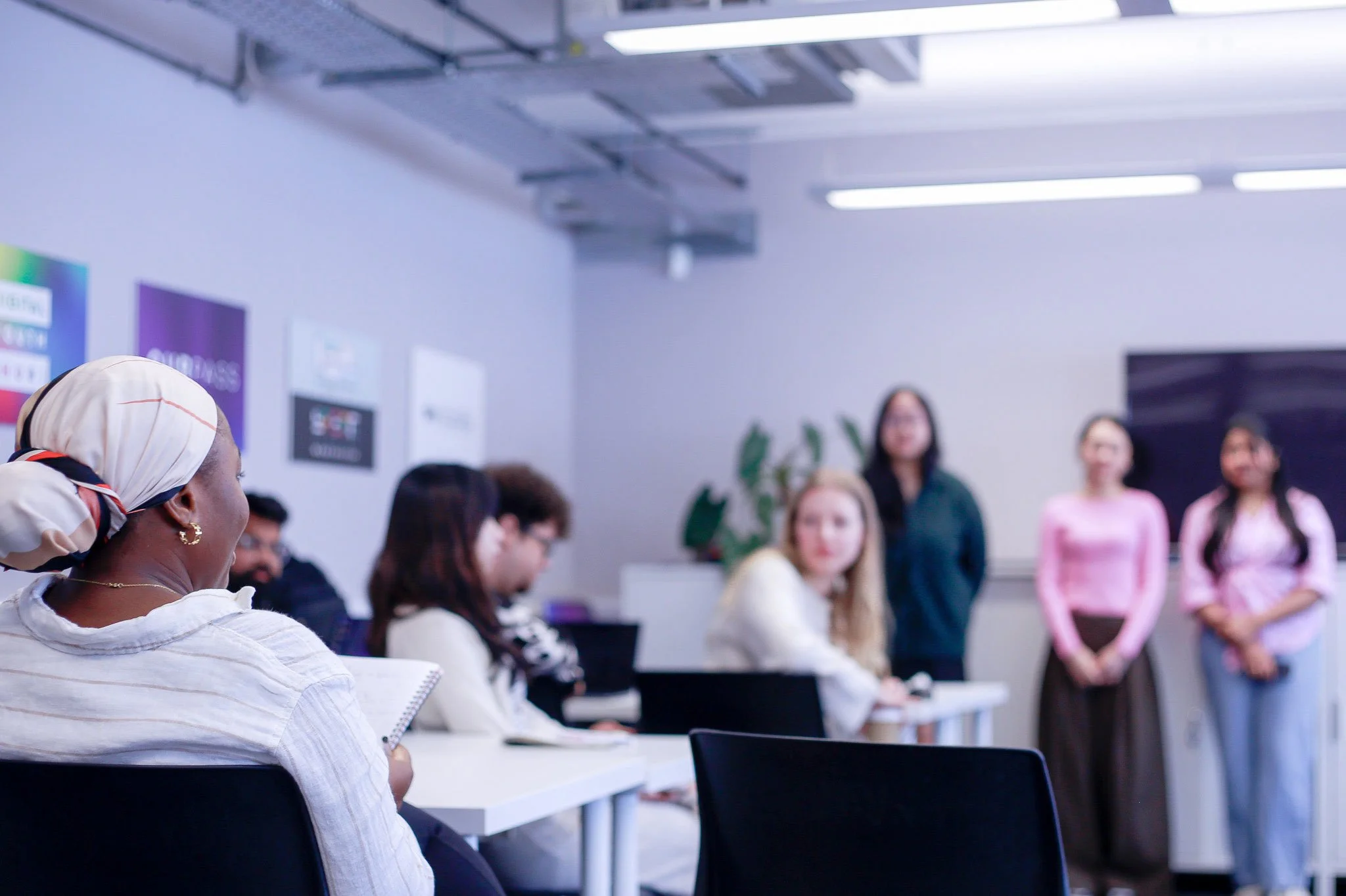 Women speaking at a meeting in a modern conference room with seated attendees, mostly women, listening attentively.
