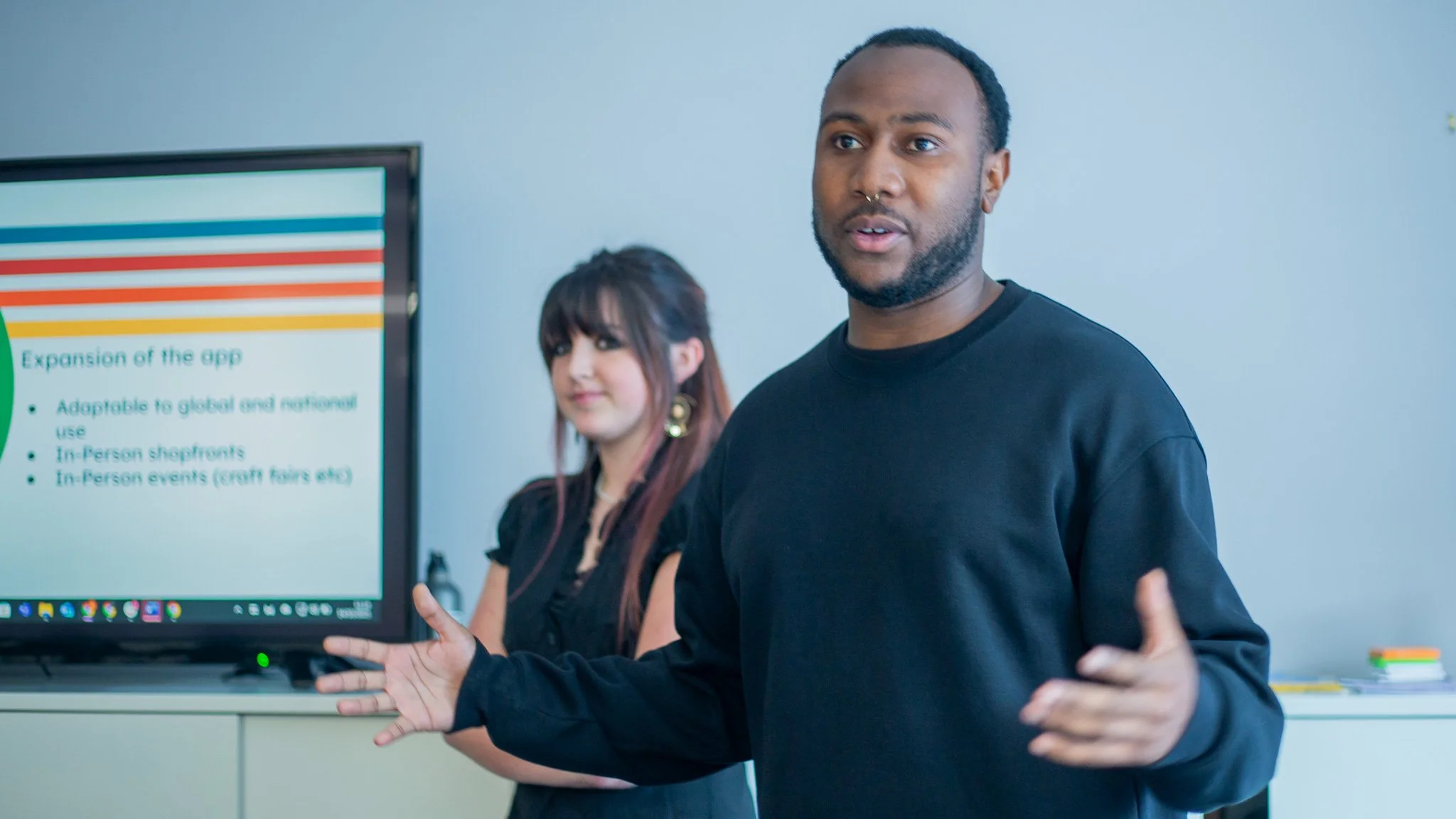 A man in a black shirt giving a presentation in front of a screen, with a woman standing behind him. The screen displays a slide titled 'Expansion of the app' with bullet points.